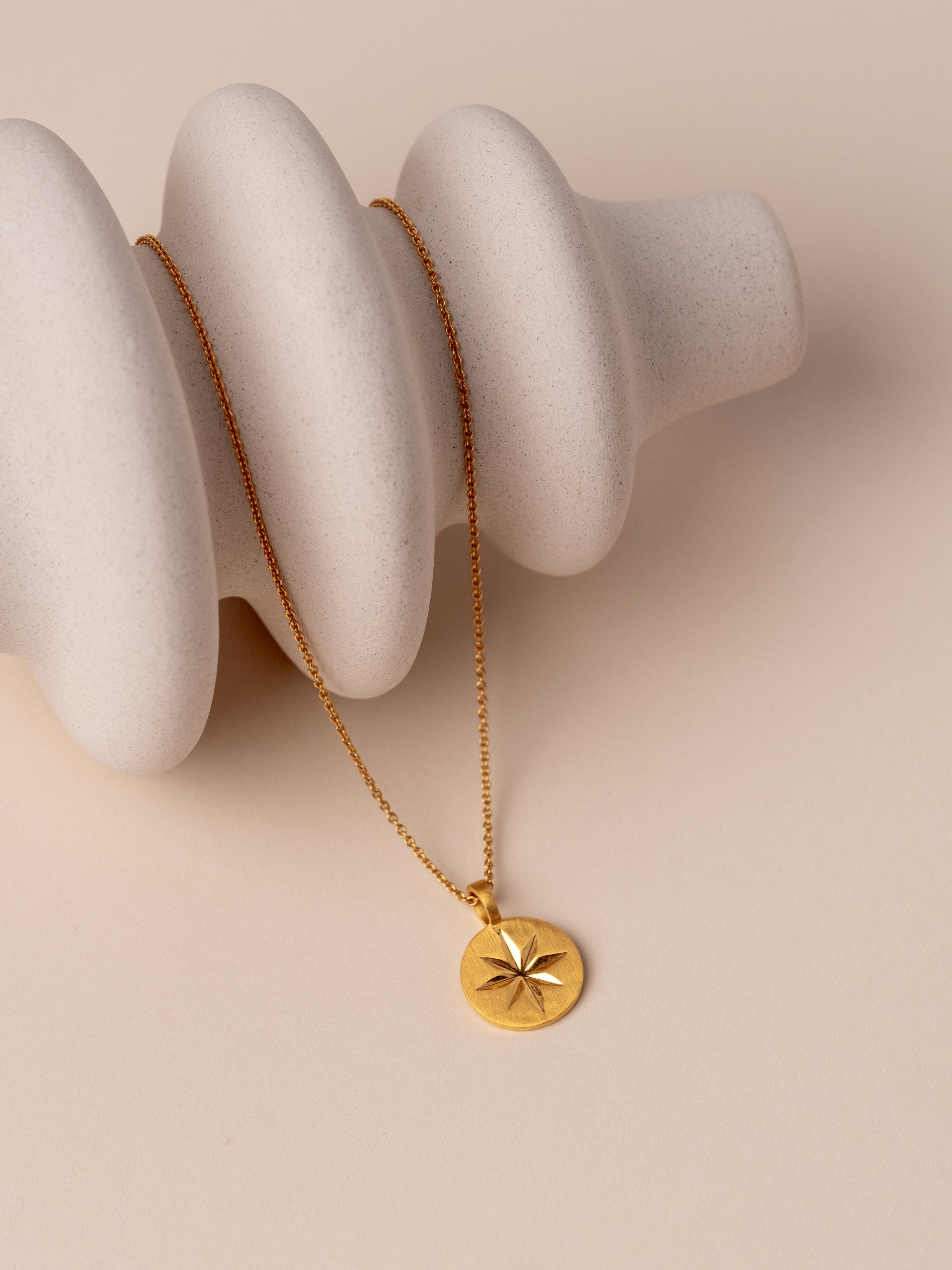 Gold necklace with a circular pendant featuring an engraved star or flower design, displayed on a textured white jewelry holder.