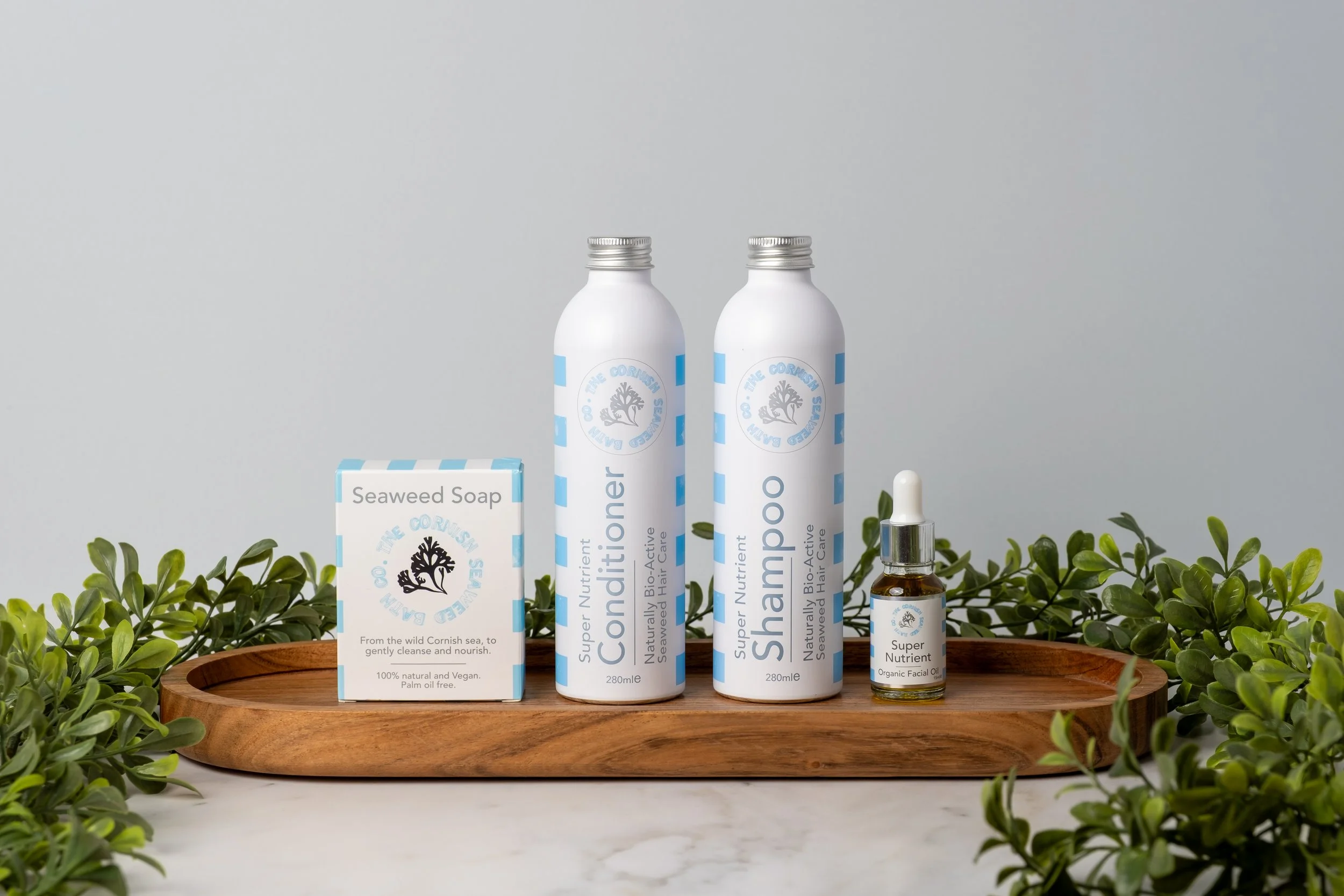 White shampoo, conditioner, serum and soap bar sitting on a wooden board on a grey/blue background with green foliage props