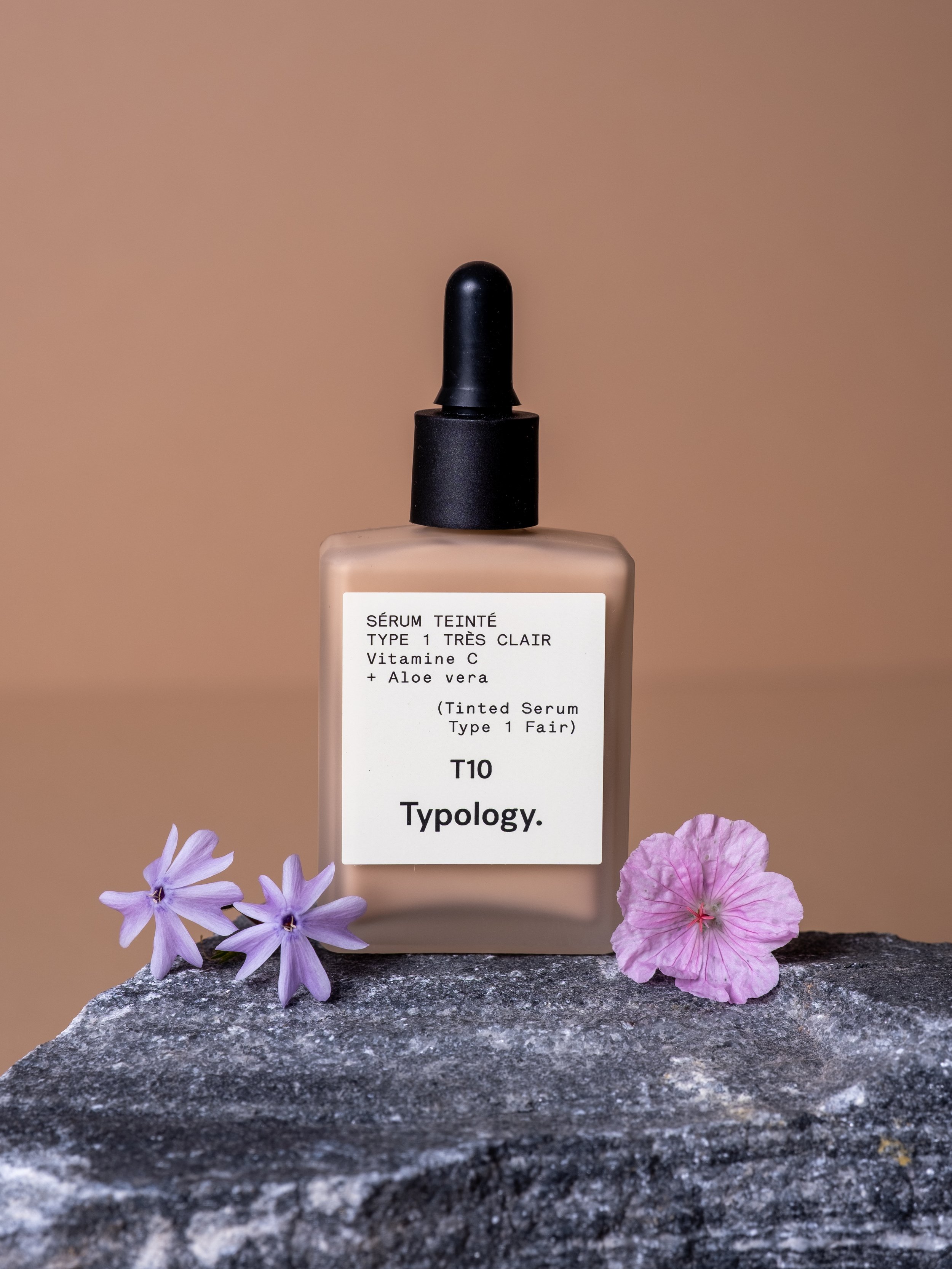 Foundation bottle sitting on a grey stone in front of a brown backdrop with pink and purples flowers each side 