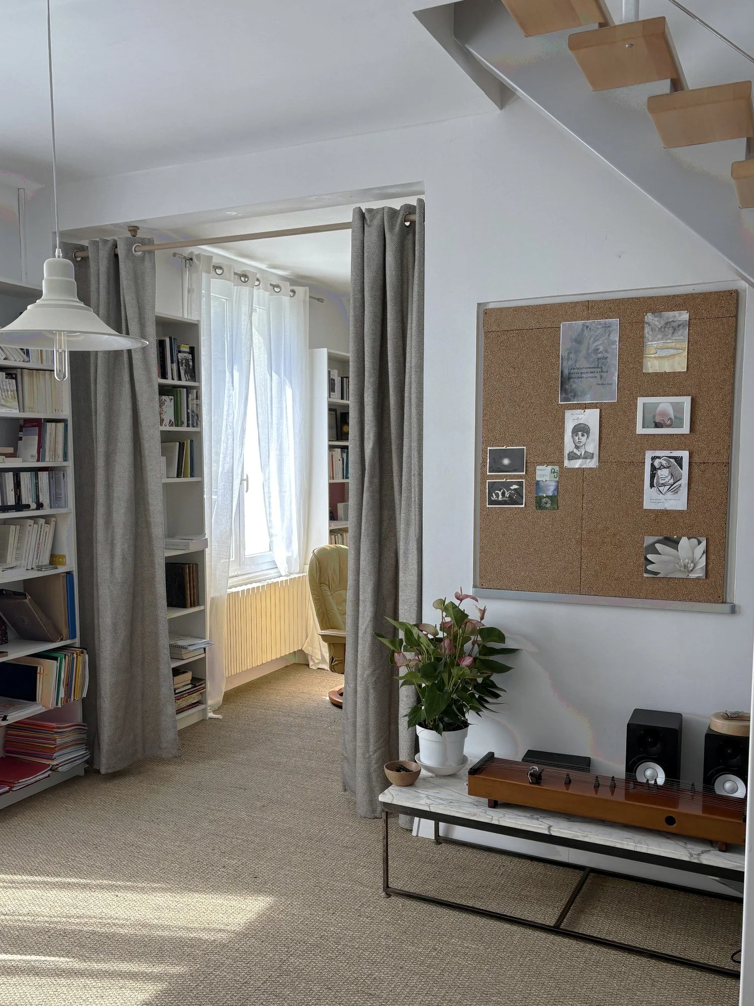 Intérieur d'une pièce lumineuse avec des étagères remplies de livres, une fenêtre laissant entrer la lumière, un fauteuil crème, une plante verte en pot, un tableau de liège avec des œuvres d'art et citations, une table basse avec un disque de vinyle, et un éclairage suspendu blanc.