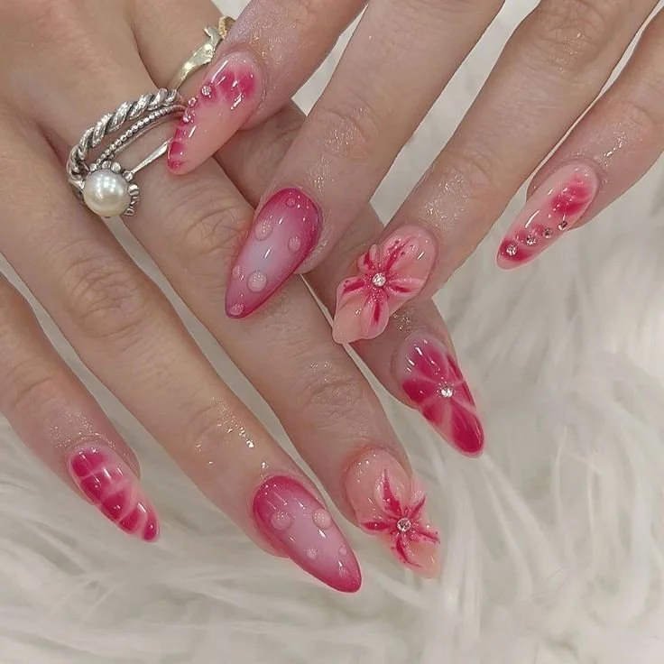Hands with red and pink floral nail art, featuring glossy polka dots and patterns, adorned with rings on white fur background.