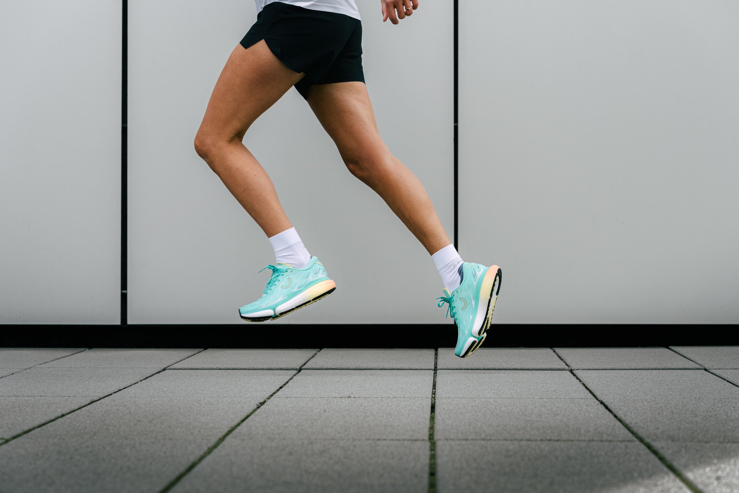 Beine einer Person in Shorts beim Laufen, mit türkisfarbenen Laufschuhen und weißen Socken, Hintergrund ist eine graue Wand.
