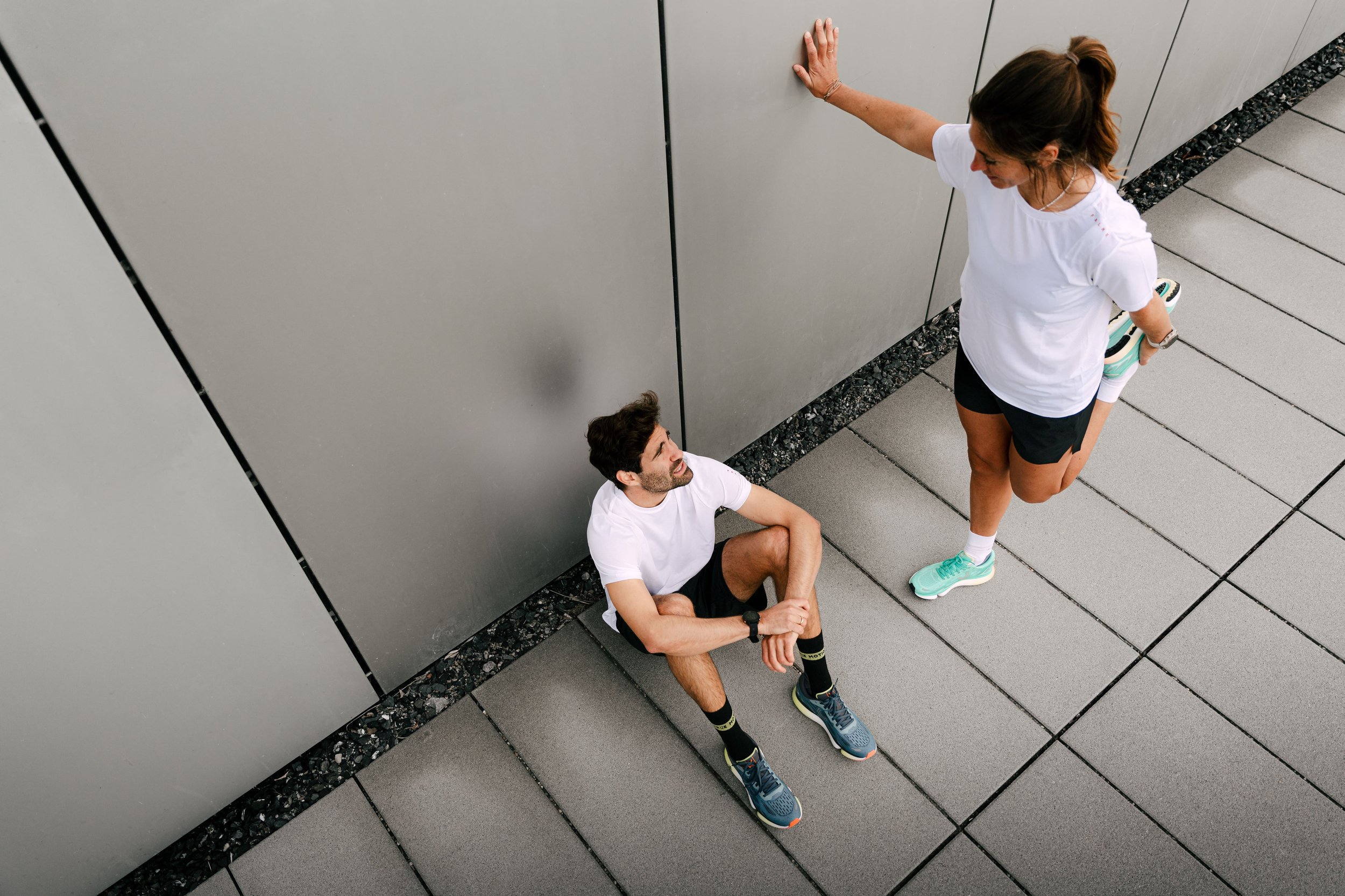 Zwei Personen in Sportkleidung, eine sitzend und die andere stehend, dehnen sich auf einem Gehweg neben einer Wand.