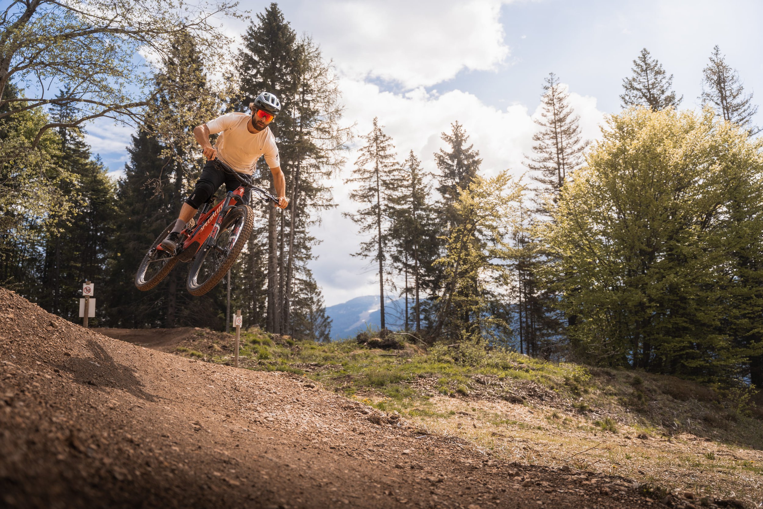 Mountainbiker springt auf einem Radweg im Wald, umgeben von Bäumen und blauem Himmel.