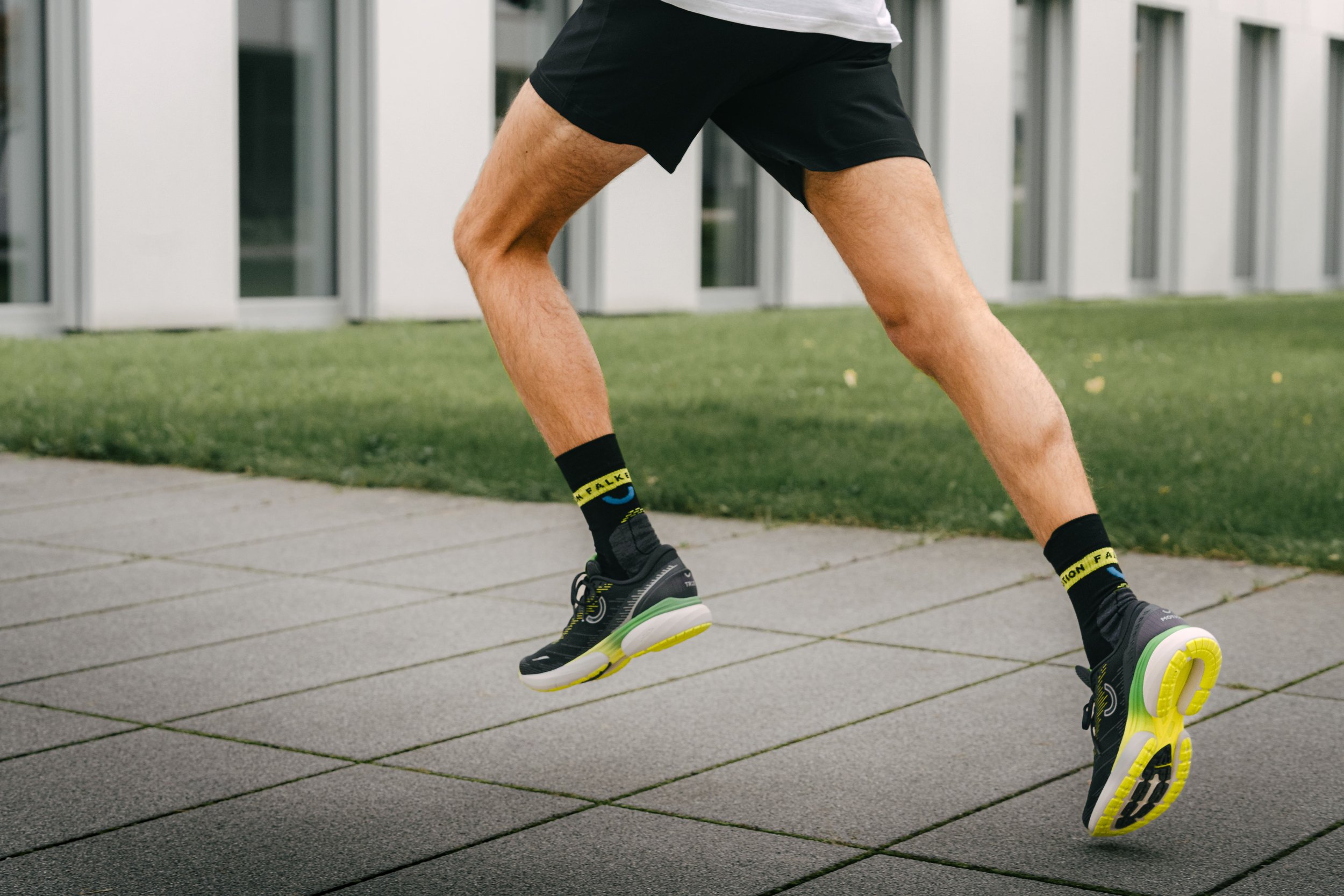 Beine eines laufenden Mannes in schwarzen Sportschuhen und kurzen Hosen auf einem gepflasterten Weg neben einer grünen Wiese.