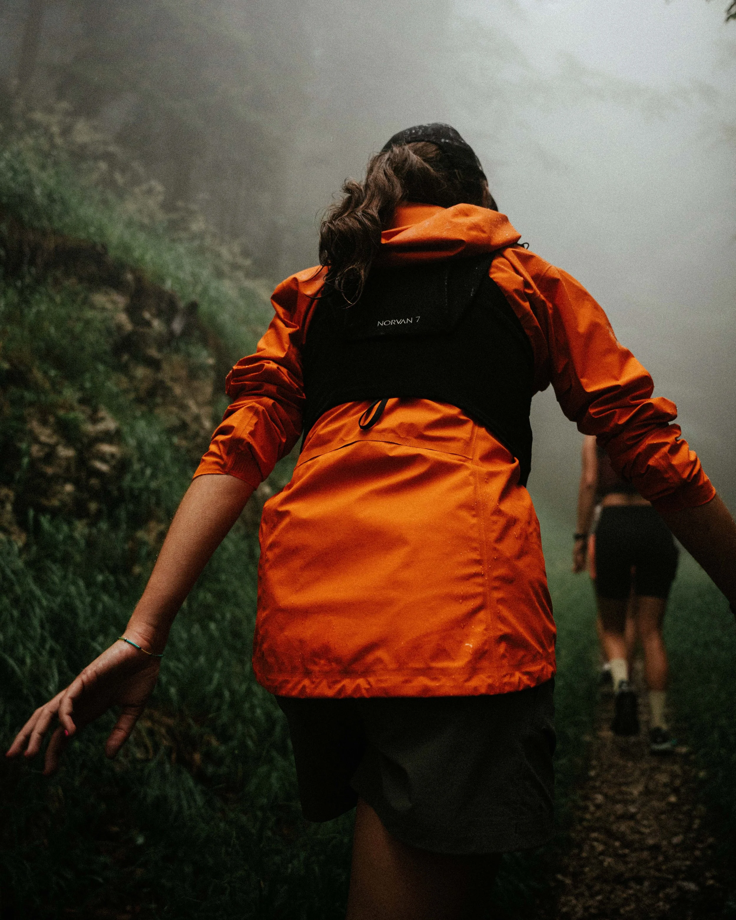Zwei Frauen beim Trailrunning auf einem nebeligen Pfad, eine trägt eine orange Jacke und einen schwarzen Rucksack.