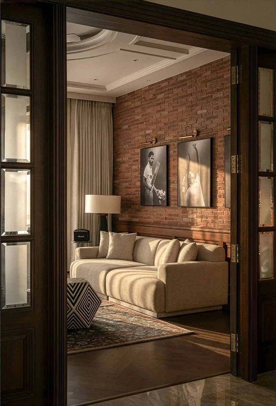 Living room with brick wall, beige sofa, black and white photographs on wall, large curtain, floor lamp, patterned rug, wooden floors, and glass door.