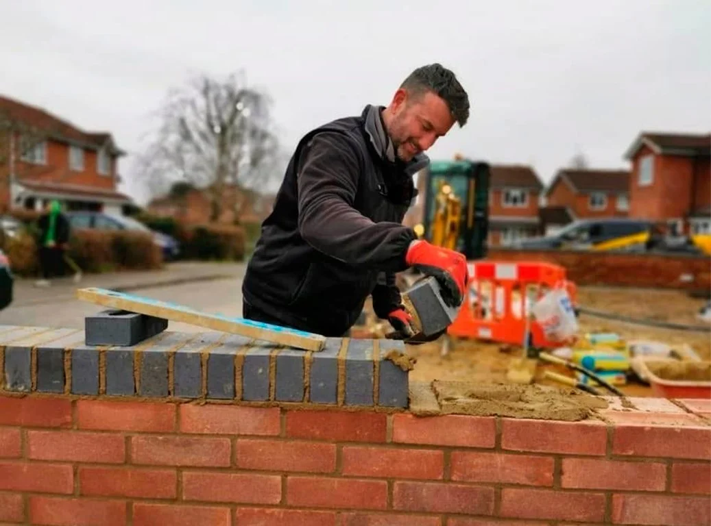 A construction worker laying bricks on an outdoor wall, wearing red gloves. A trowel and mortar are visible. Background shows houses and a construction site with equipment.
