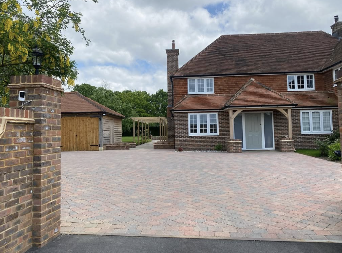 Brick house with a large driveway, wooden garage, and a pergola in a yard setting.