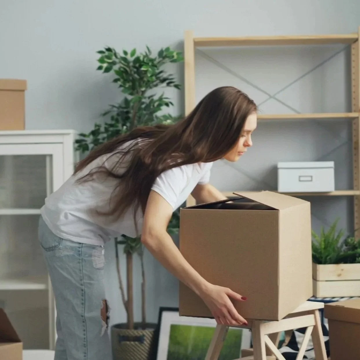 A woman packing a cardboard box in a living room with other boxes and houseplants.