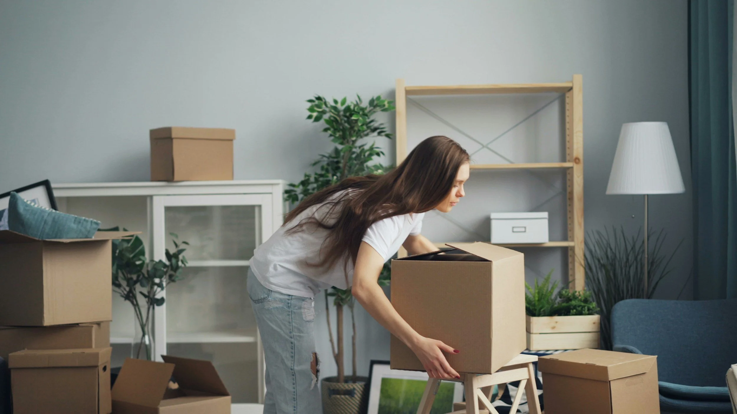 A woman packing a cardboard box in a living room with other boxes and houseplants.