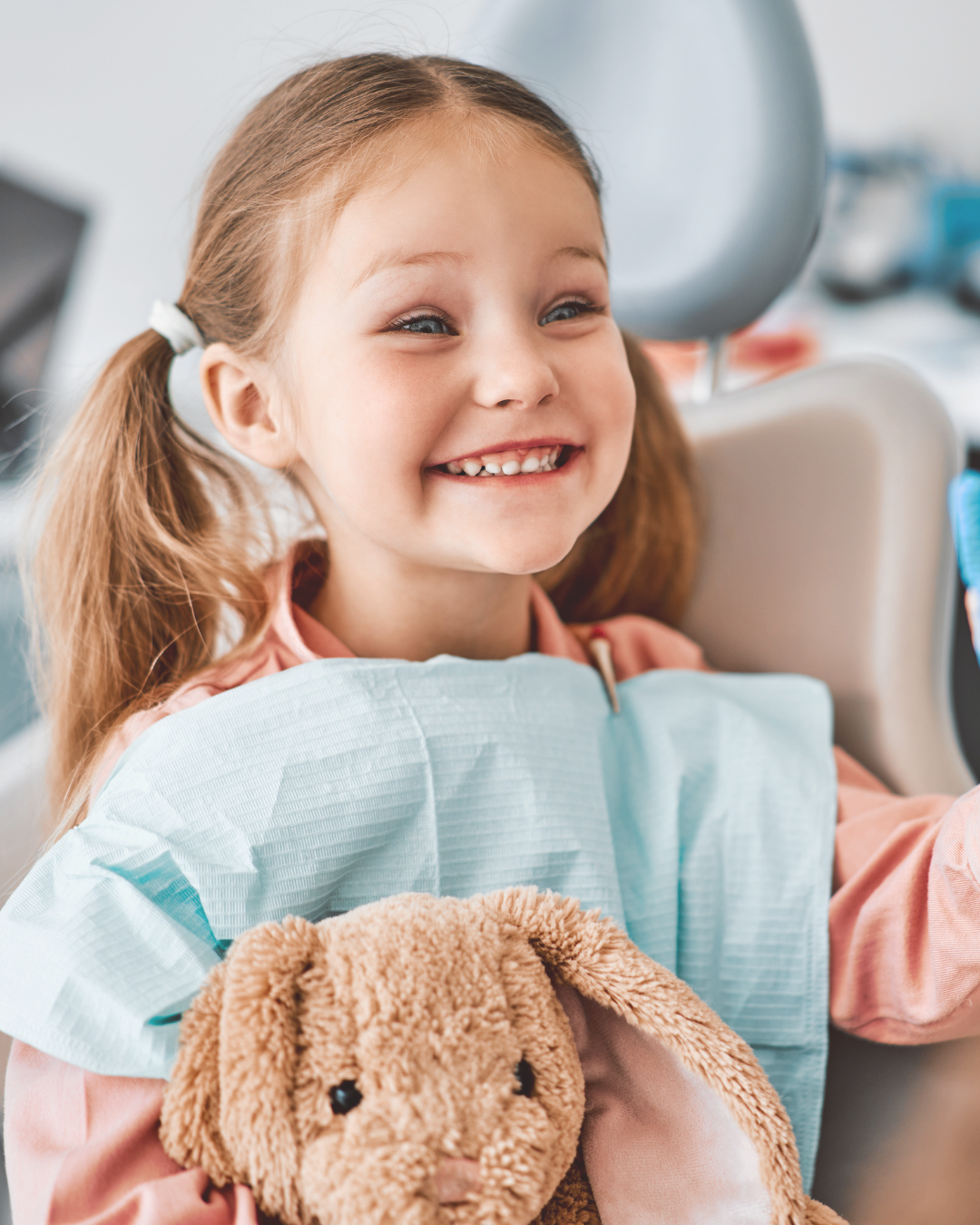 A young girl with red hair in pigtails smiling at a dental appointment, holding a brown stuffed bunny, wearing a dental gown, in a dental clinic.