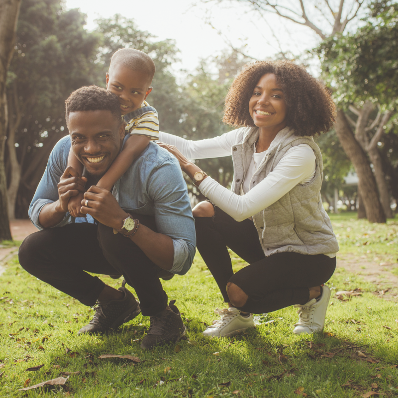 A happy family of three, an African American man, woman, and young boy, playing together outdoors in a park with green trees and grass, smiling and enjoying their time.