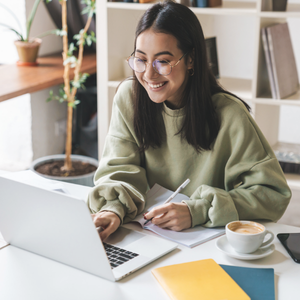 Young woman with glasses smiling while working on her laptop at a desk with a notebook, coffee cup, and yellow folder