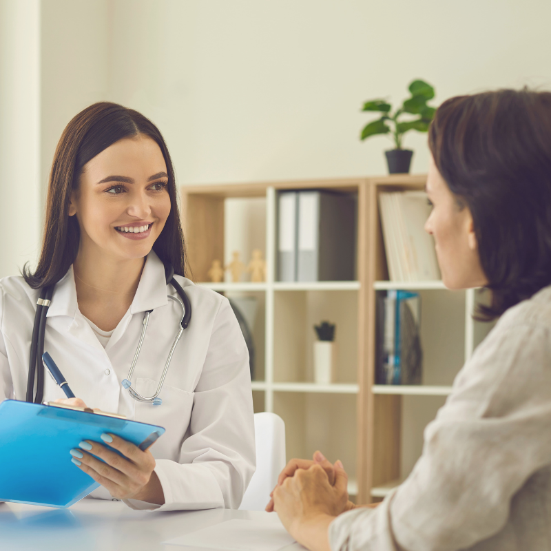 A female doctor smiling and holding a clipboard while talking to a female patient in an office setting.