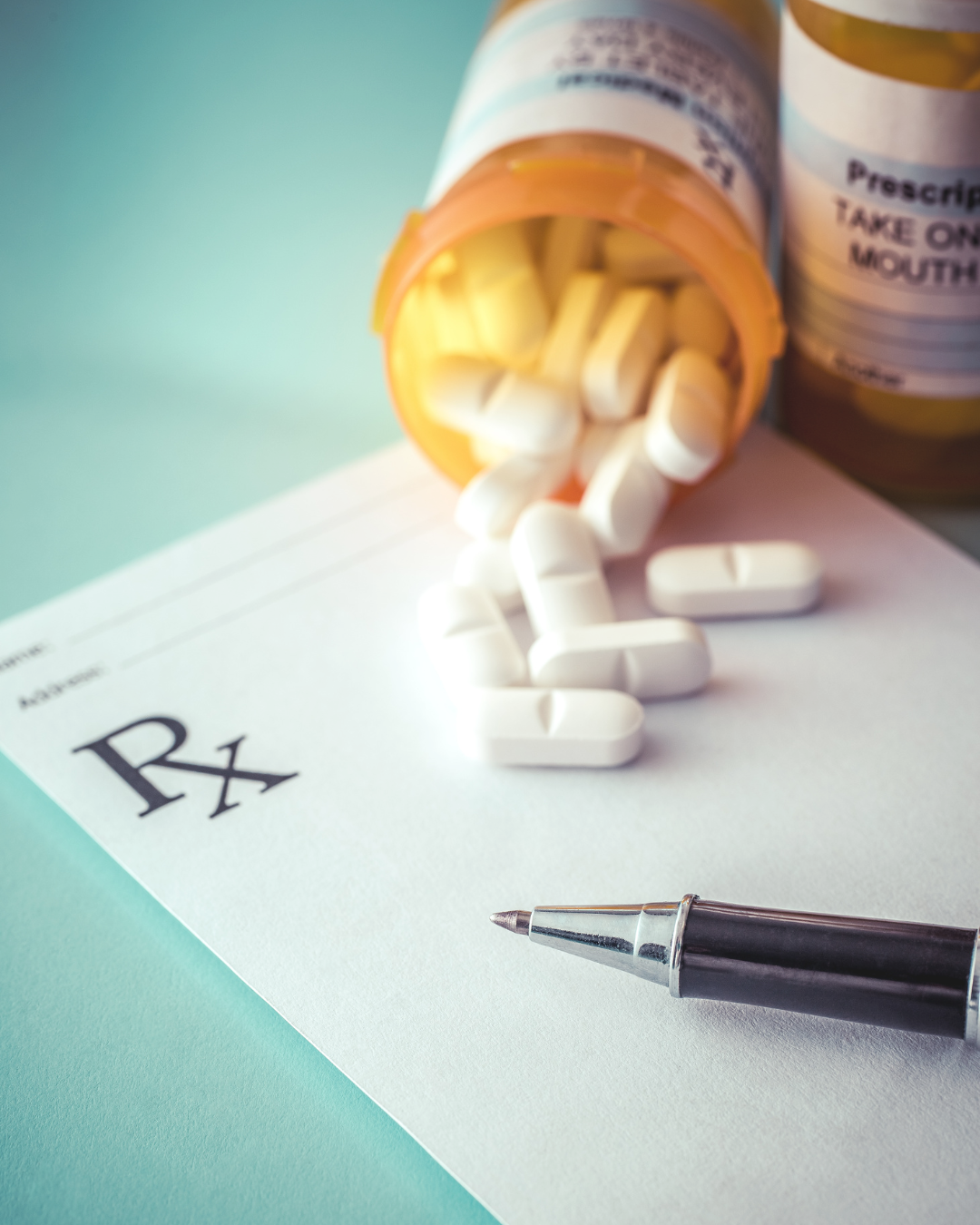 An orange prescription pill bottle spilling white pills onto a pharmacy prescription pad and a silver pen.
