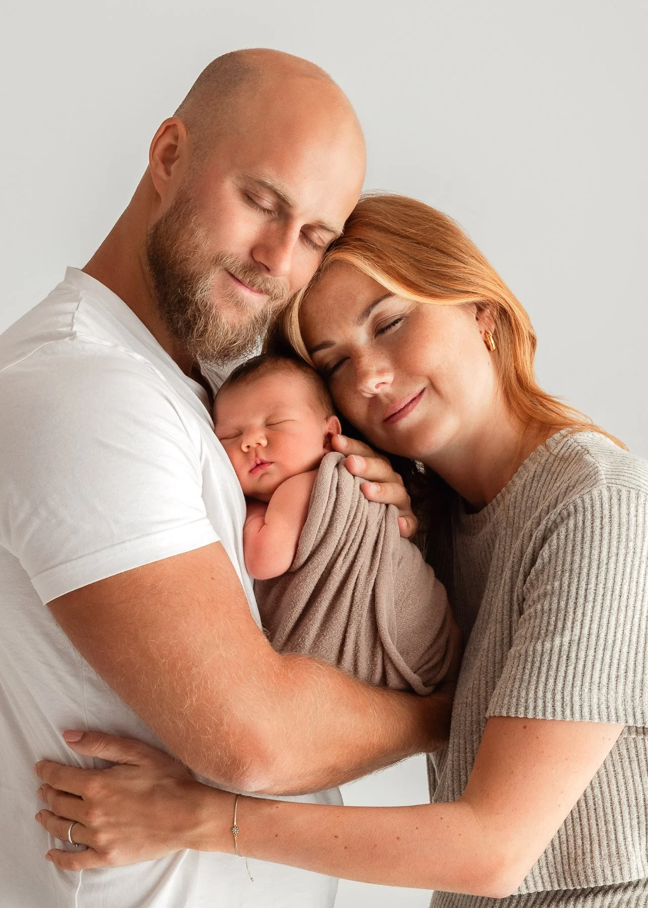 Ein glückliches Elternpaar hält ein schlafendes Baby in den Armen, beide haben ihre Augen geschlossen und lächeln sanft, vor einem weißen Hintergrund.