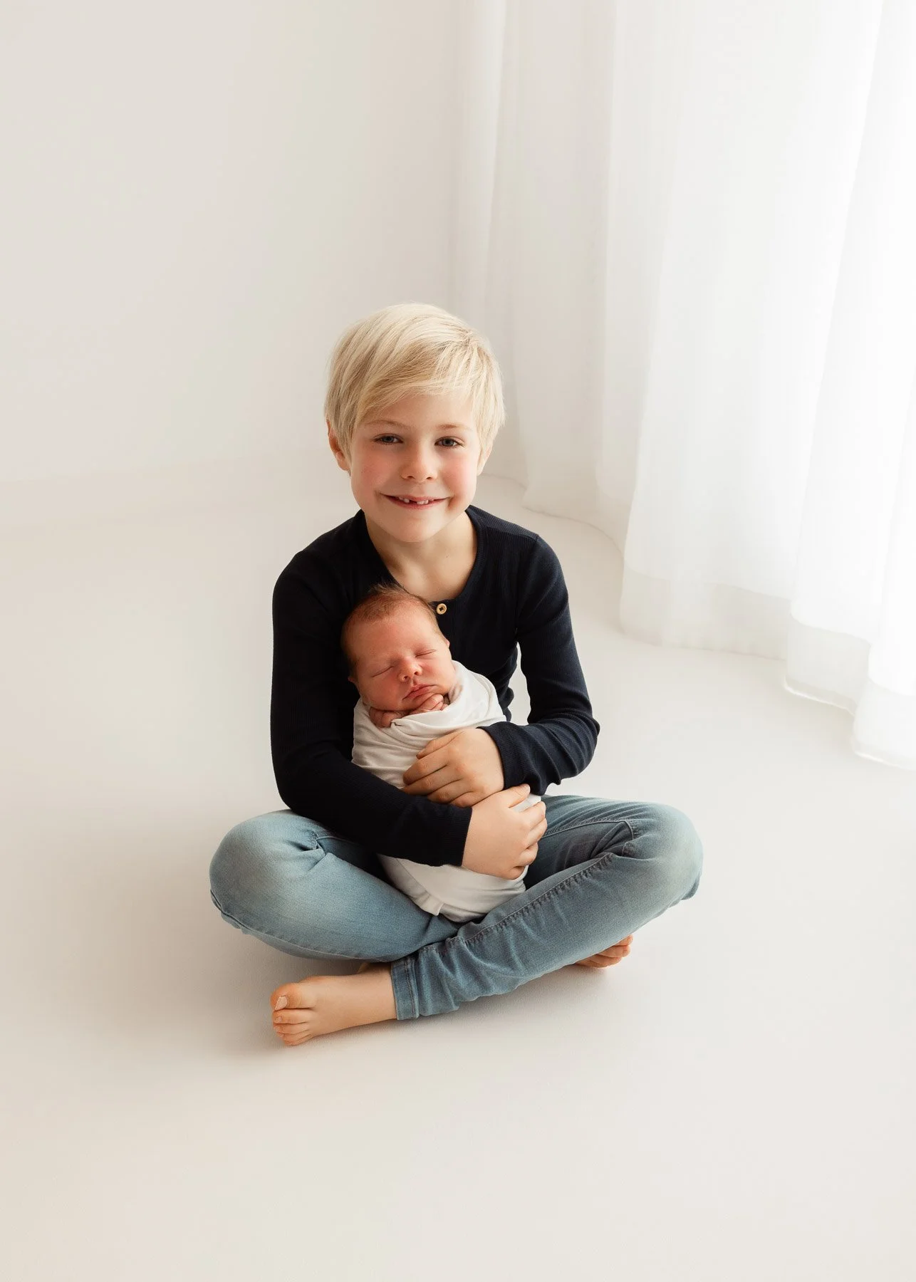 Ein älterer Junge sitzt im Schneidersitz auf dem Boden und hält ein schlafendes Baby in den Armen, beide vor einem weißen Hintergrund mit weißen Vorhängen im Hintergrund.