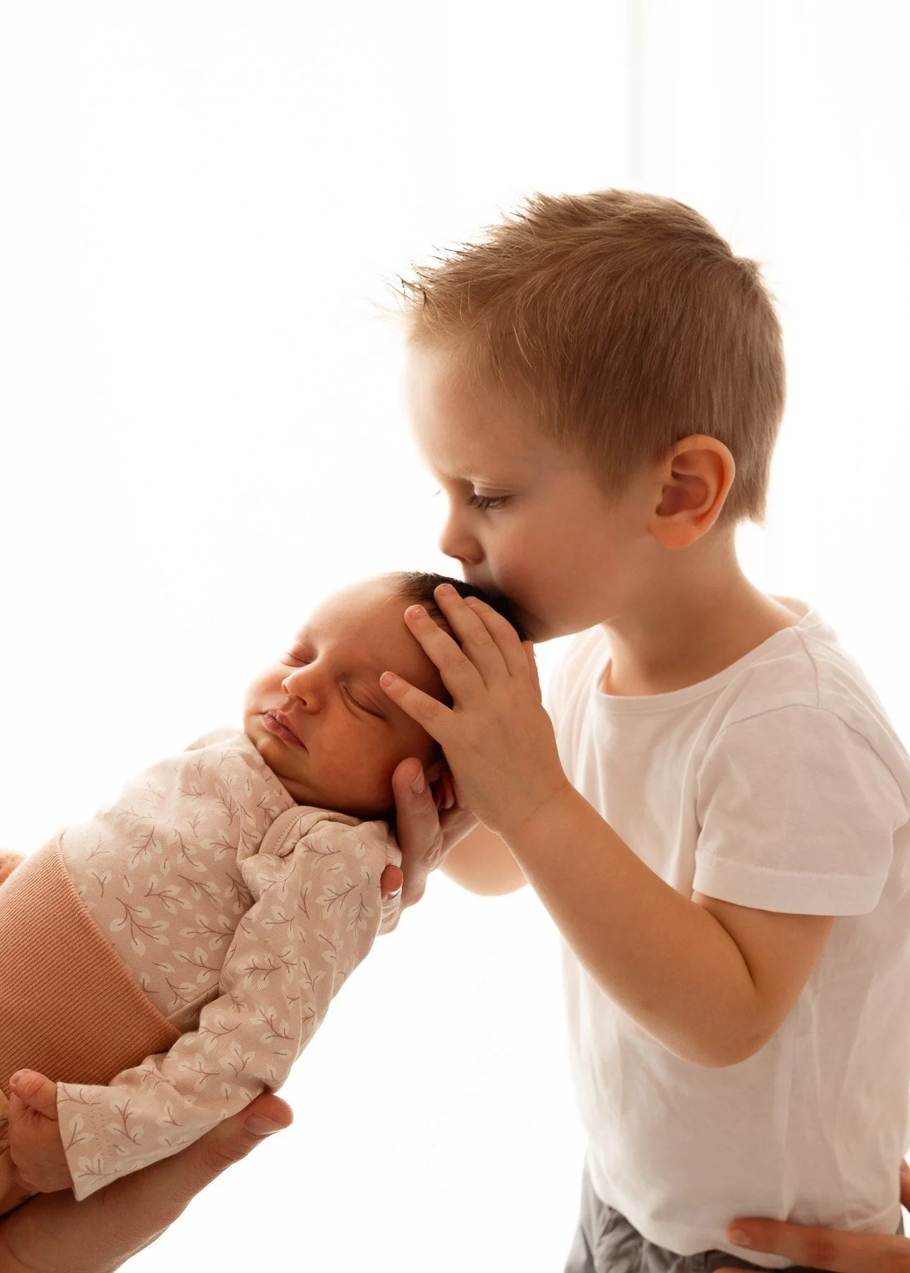 Ein kleines Kind küsst liebevoll ein Baby auf den Kopf, während es von einem Erwachsenen gehalten wird.