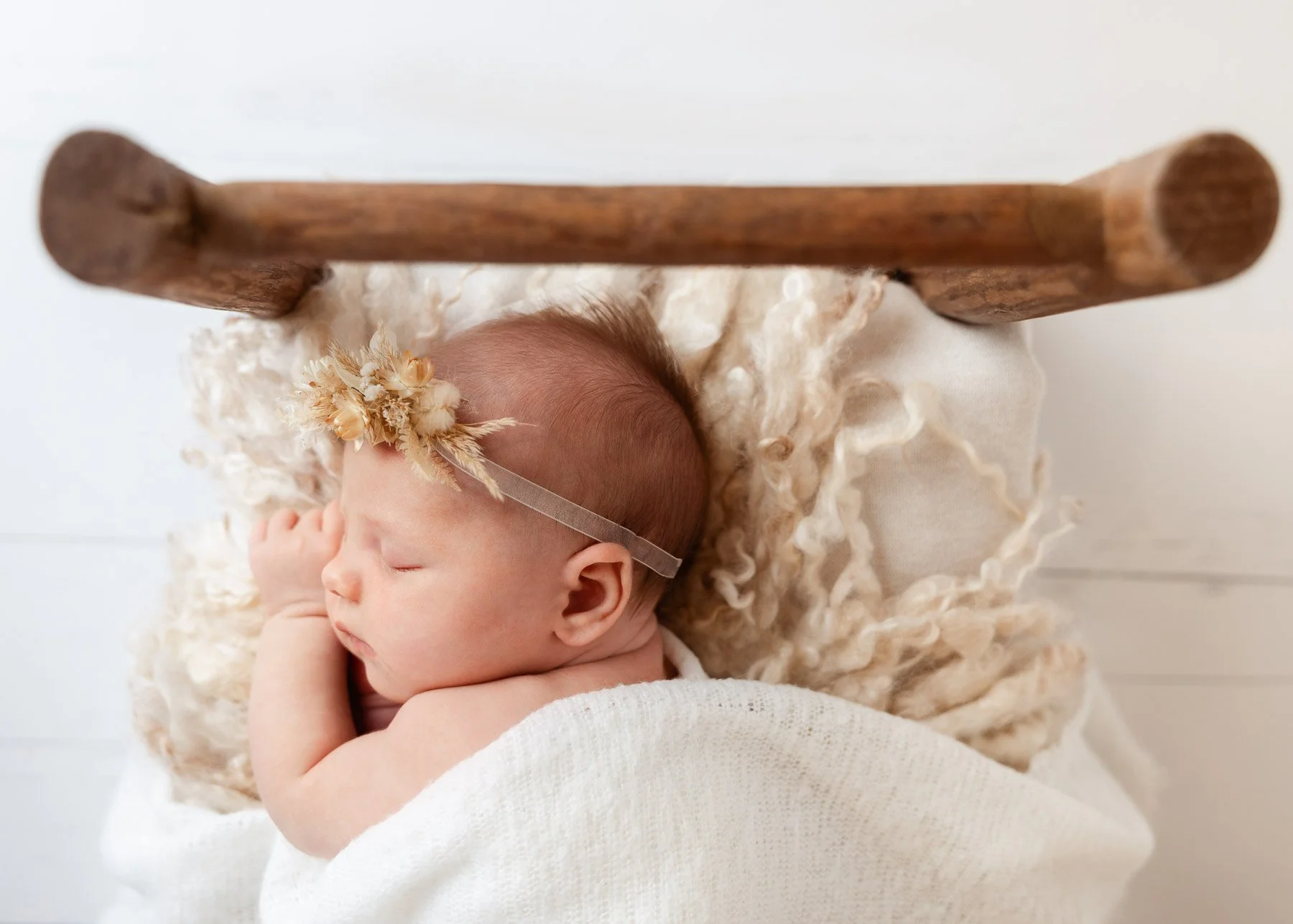 Ein schlafender Baby mit einer Blumenkrone und einem dünnen Kopfband liegt auf einem weichen, cremefarbenen Kissen in einem Korb.