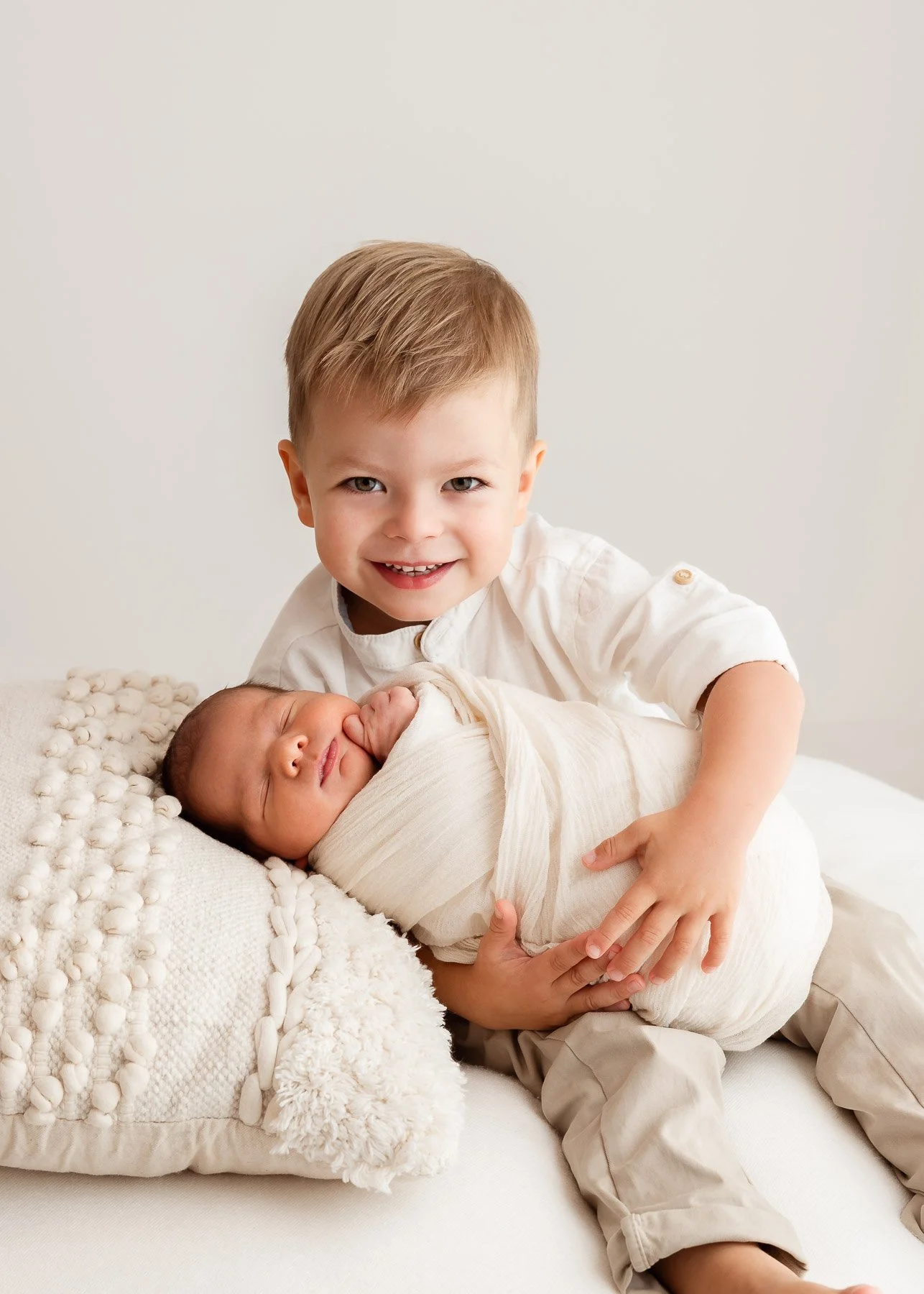 Ein freundlicher älterer Junge hält einen schlafenden neugeborenen Säugling, beide sitzen auf einem weißen Bett, die Szene ist hell und minimalistisch.
