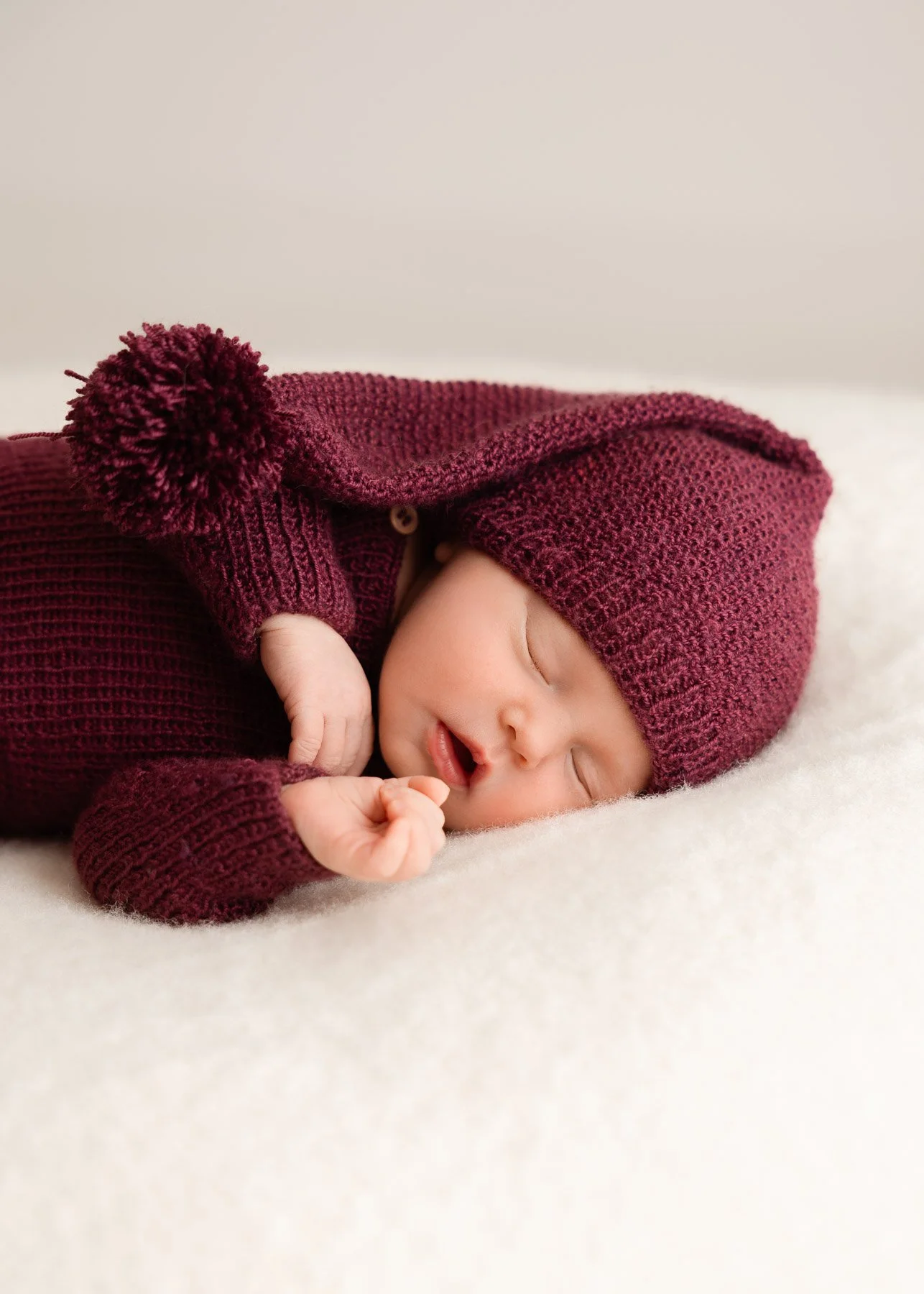 Ein schlafendes Baby, das in einem maroonfarbenen Hand-knit Outfit mit Mütze und Pompon liegt auf einem weichen weißen Tuch.