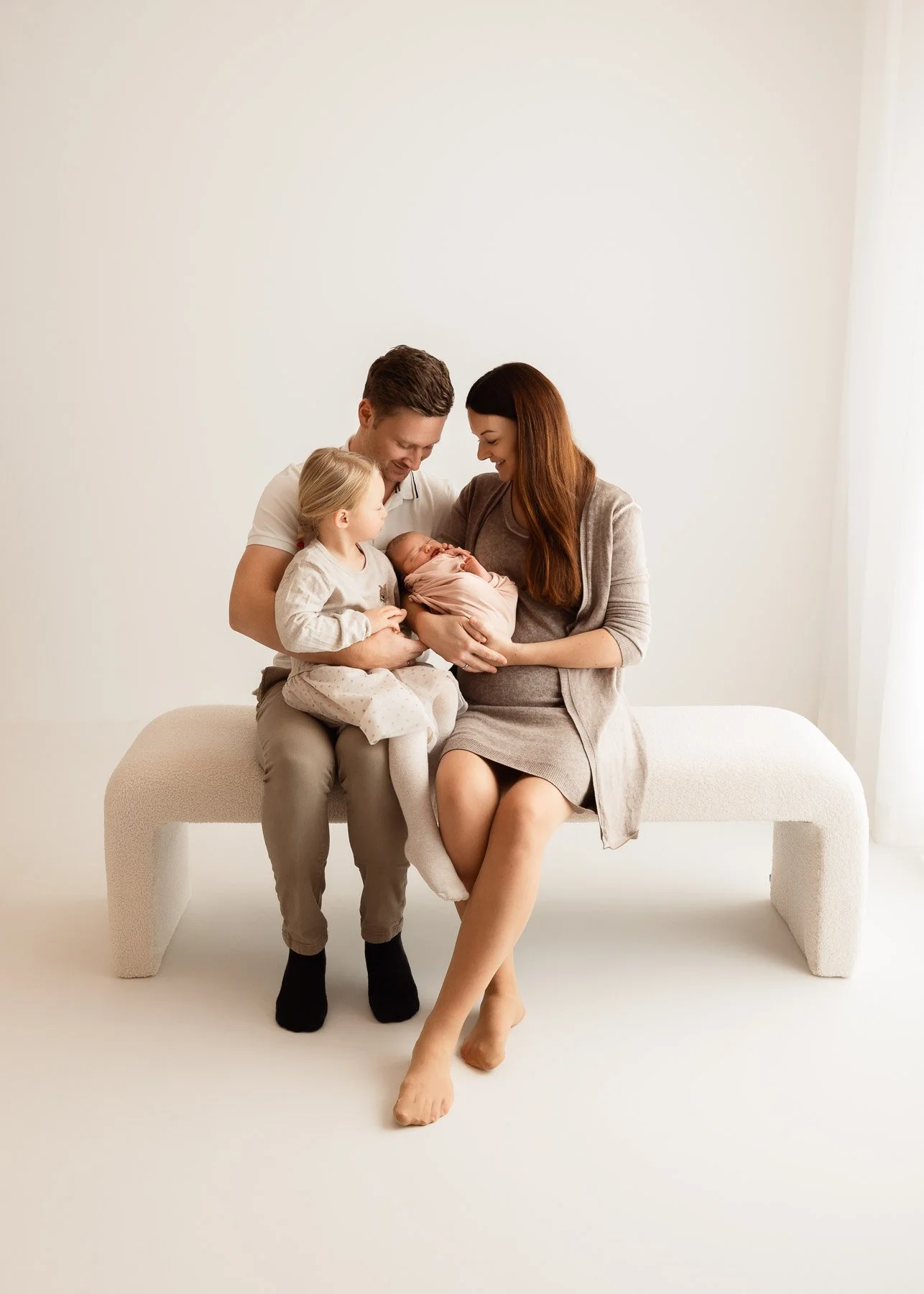Eine Familie mit einem Vater, einer Mutter, einem kleinen Kind und einem neugeborenen Baby auf einer Bank in einem hellen Raum.
