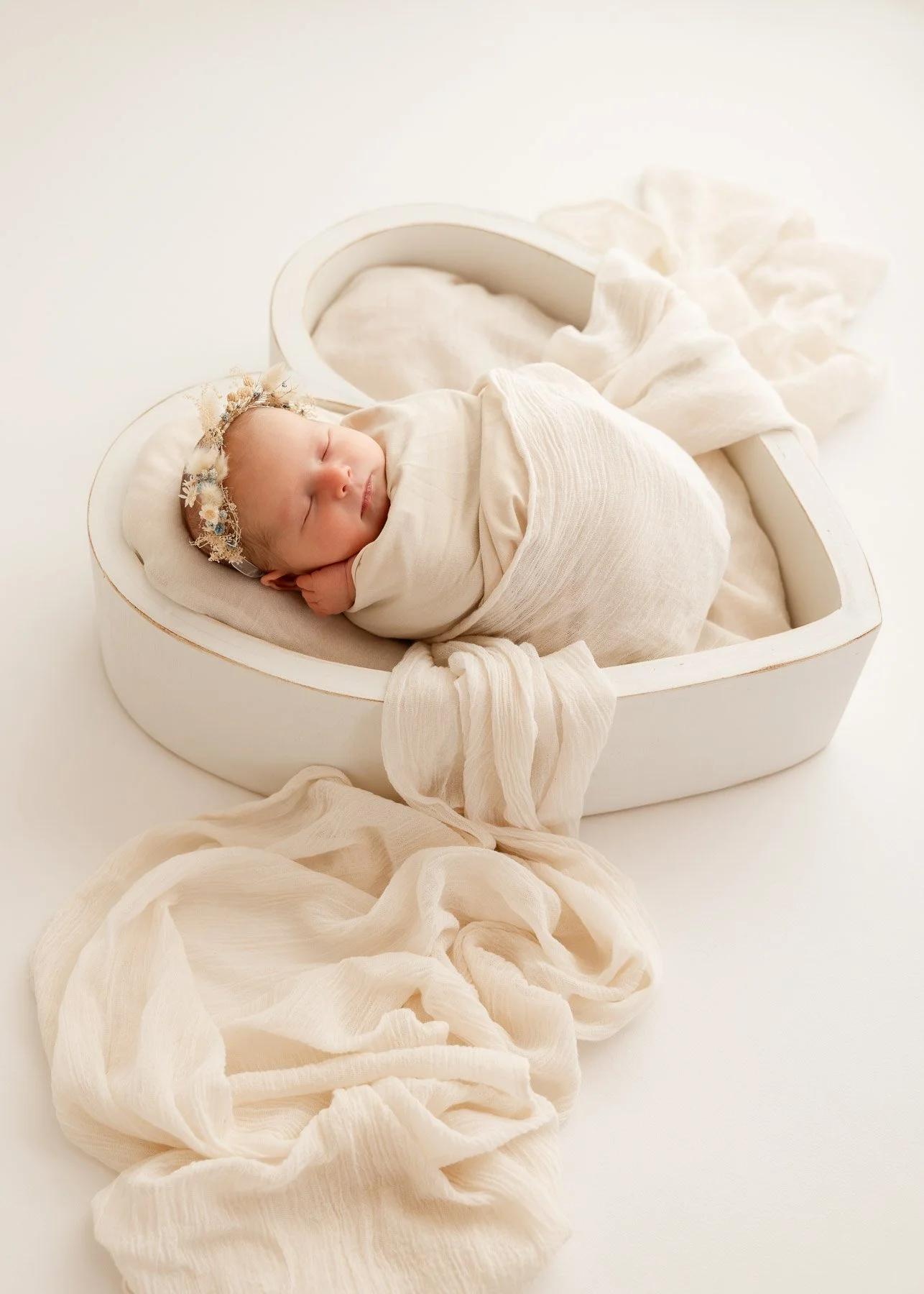 Ein schlafender neugeborener Baby in einer weißen Herz-Form-Box, umgeben von heller, lockerer Stoff, mit einem Blumenkranz am Kopf.