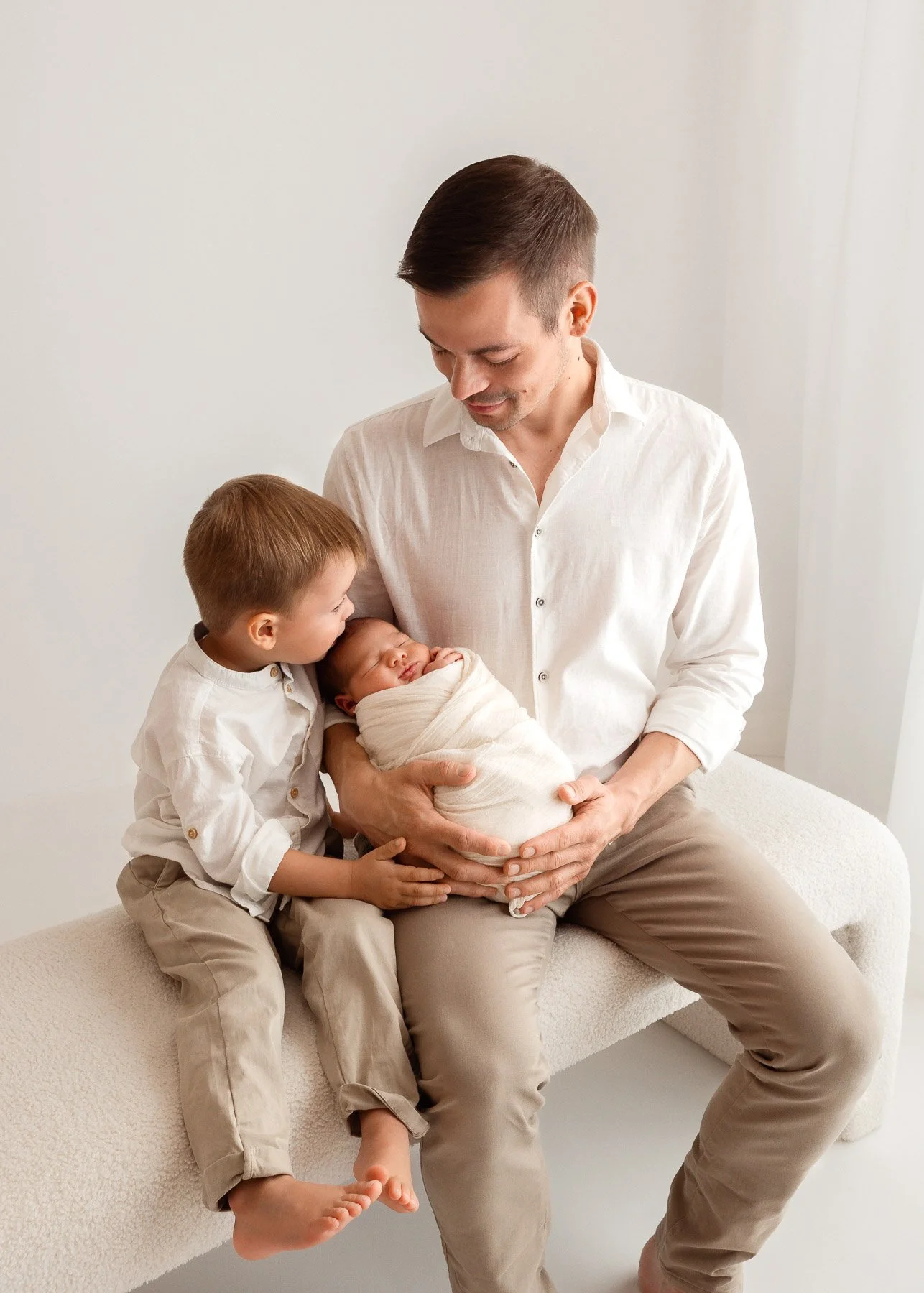 Vater hält neugeborenes Baby, das von älterem Kind geküsst wird, in einem hellen Raum.
