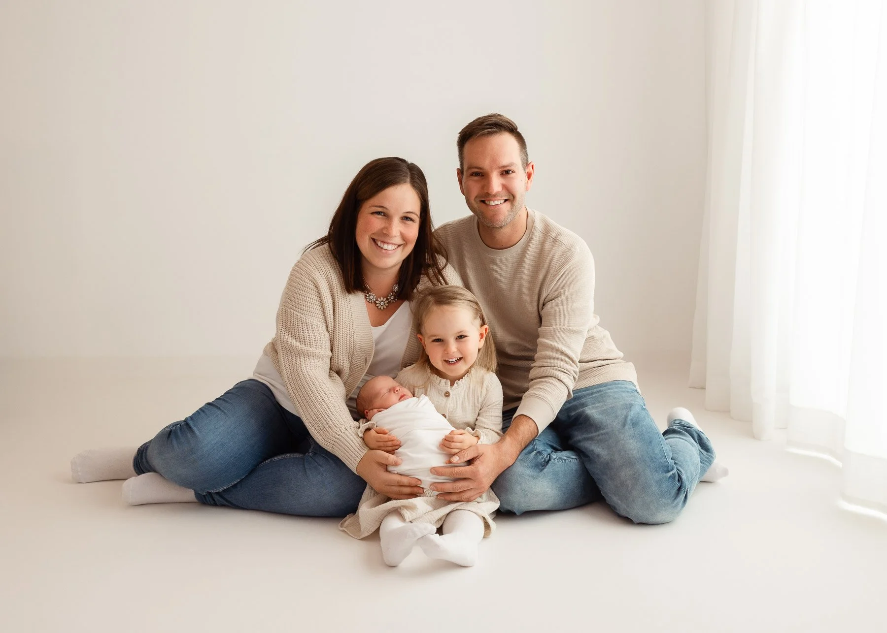 Familie mit zwei Erwachsenen, einem kleinen Mädchen und einem neugeborenen Baby sitzt auf dem Boden in einem hellen Raum und lächelt.