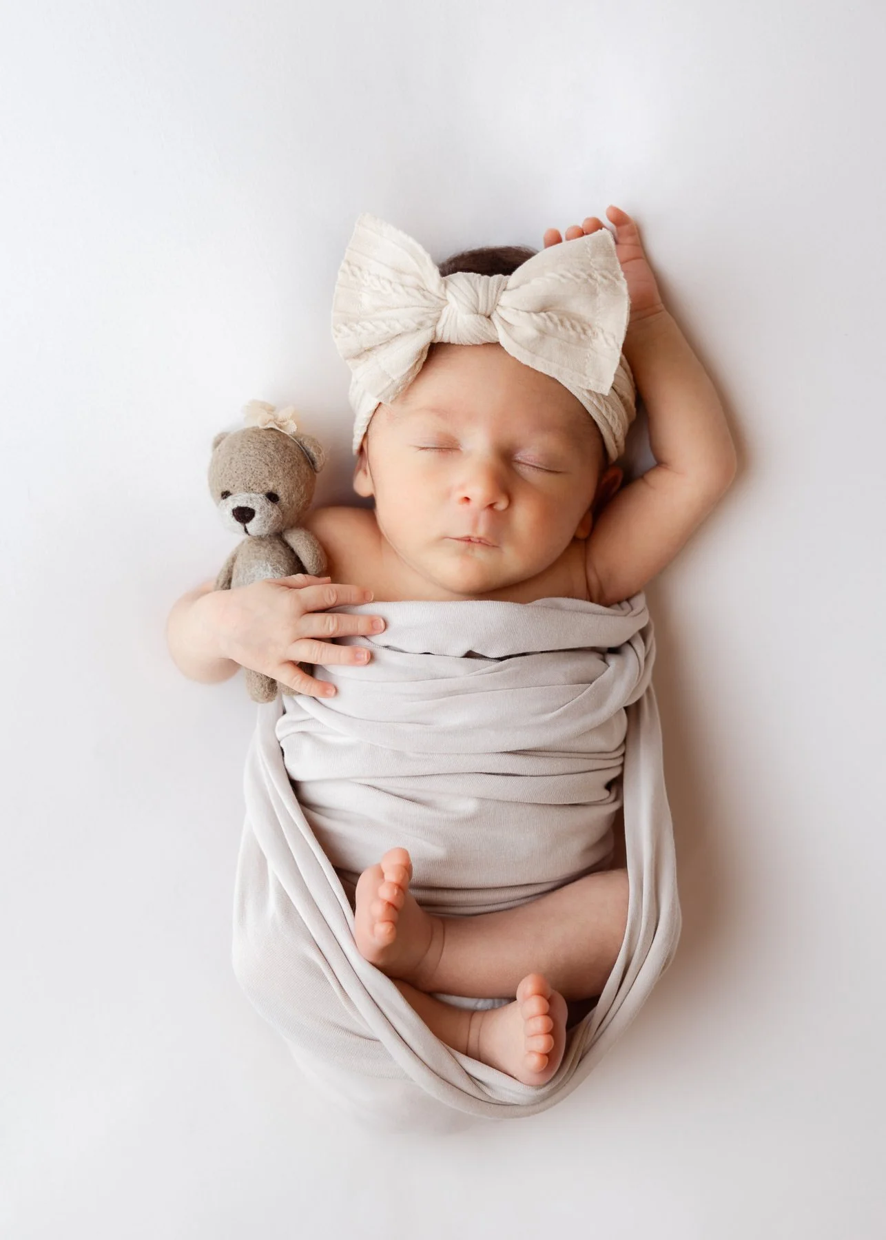 Ein schlafendes Baby mit Schleife auf dem Kopf, eingewickelt in eine weiße Decke, mit einem kleinen Stoffbären, auf weißem Hintergrund.