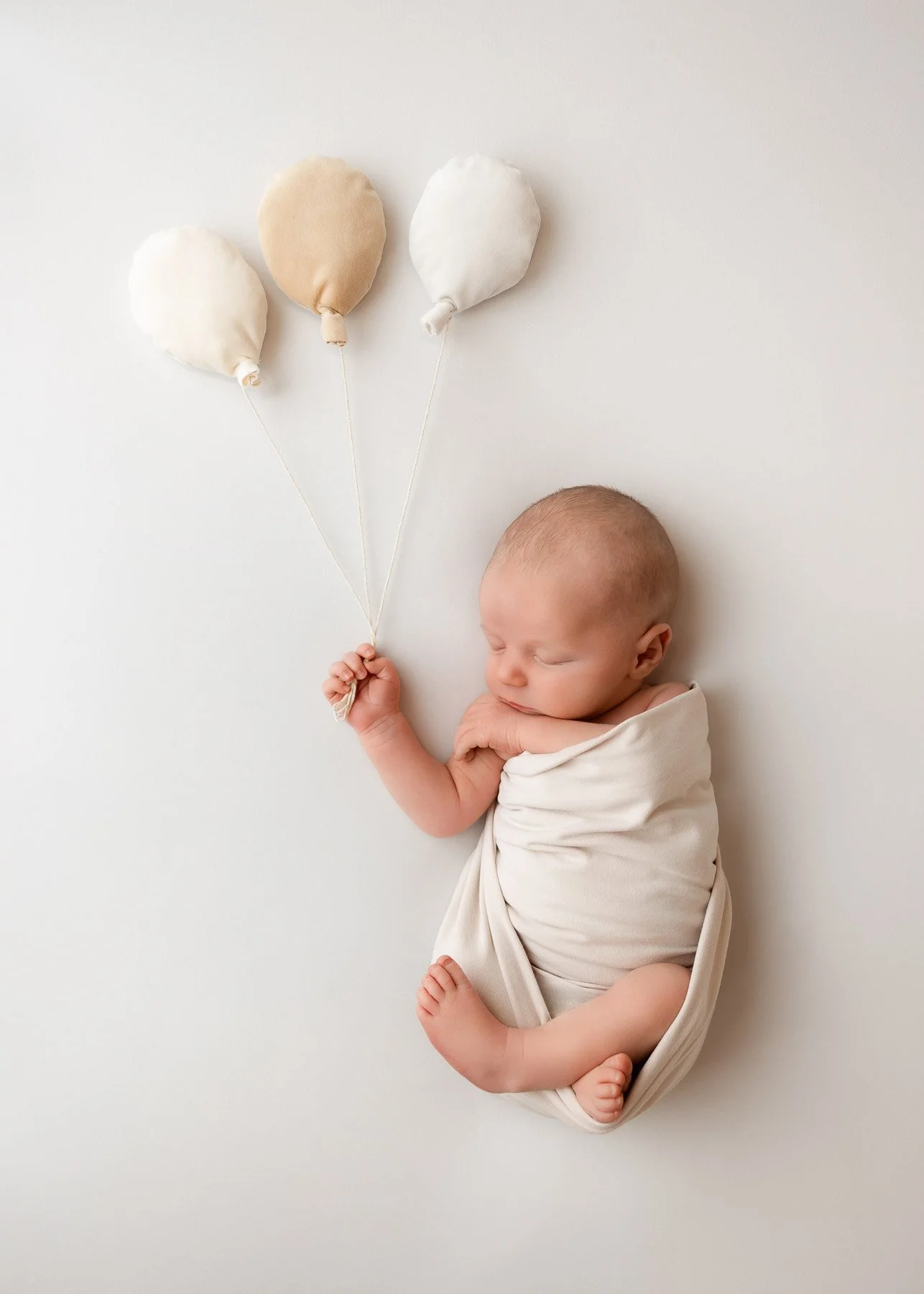 Ein schlafender Säugling, der in einem cremefarbenen Tuch gewickelt ist und auf einem weißen Hintergrund liegt, hält drei Stoffballons in Pastellfarben.