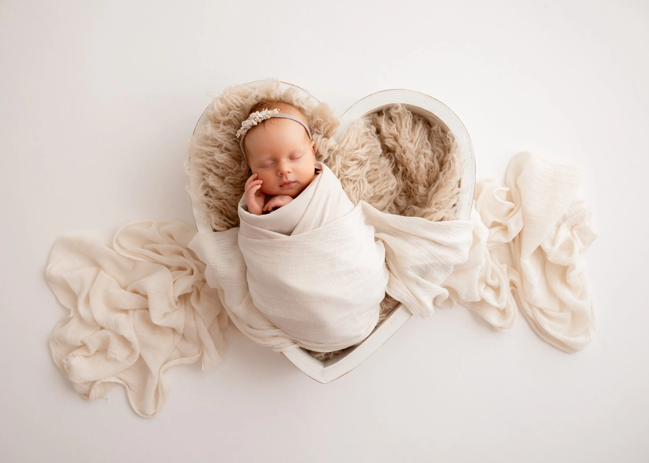 Ein schlafendes Baby in einem Herzförmigen Korb, umgeben von cremefarbenen Stoffen.