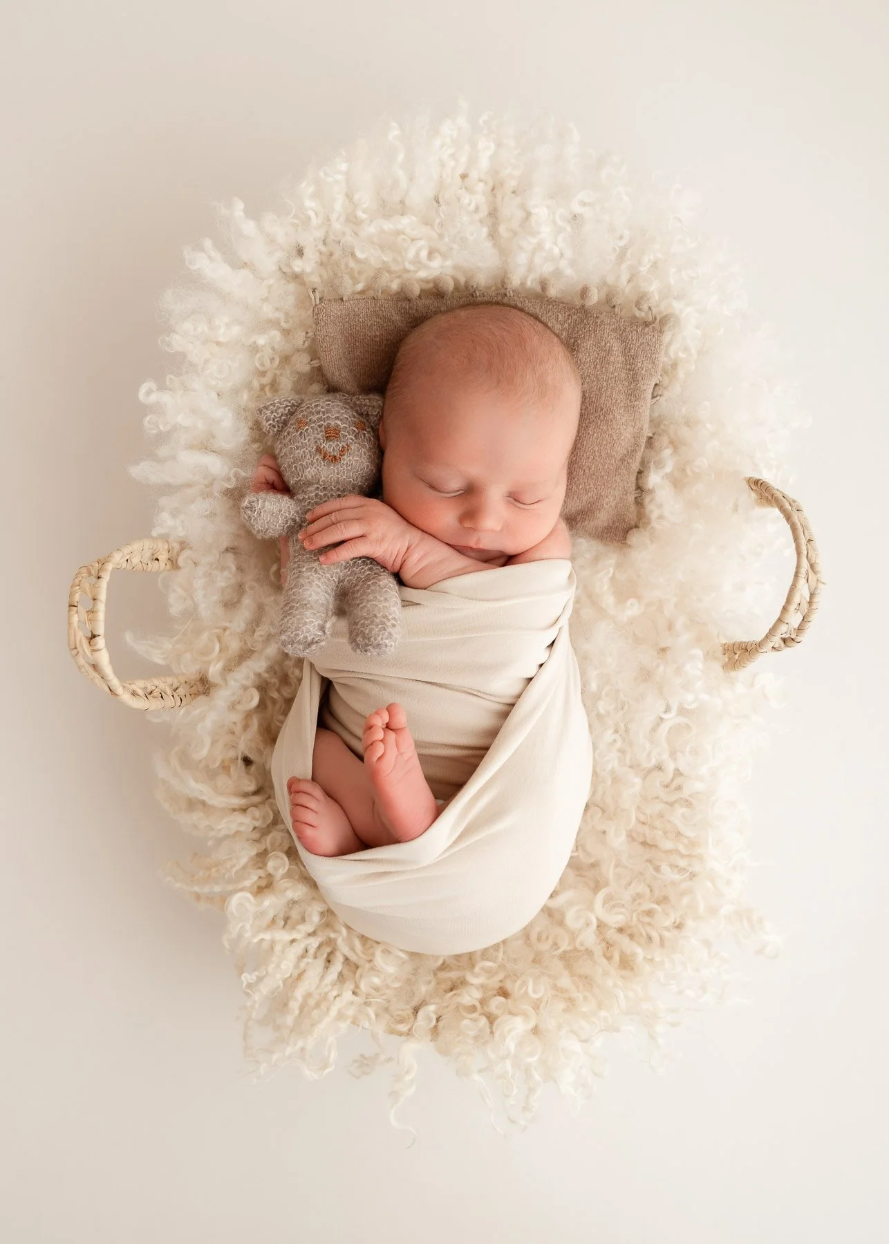 Säugling, der schläft, in einem weißen Tuch eingewickelt, auf einem weichen, cremefarbenen Fell in einem Korb, mit einem kleinen grauen Stoffhund aus Wolle.