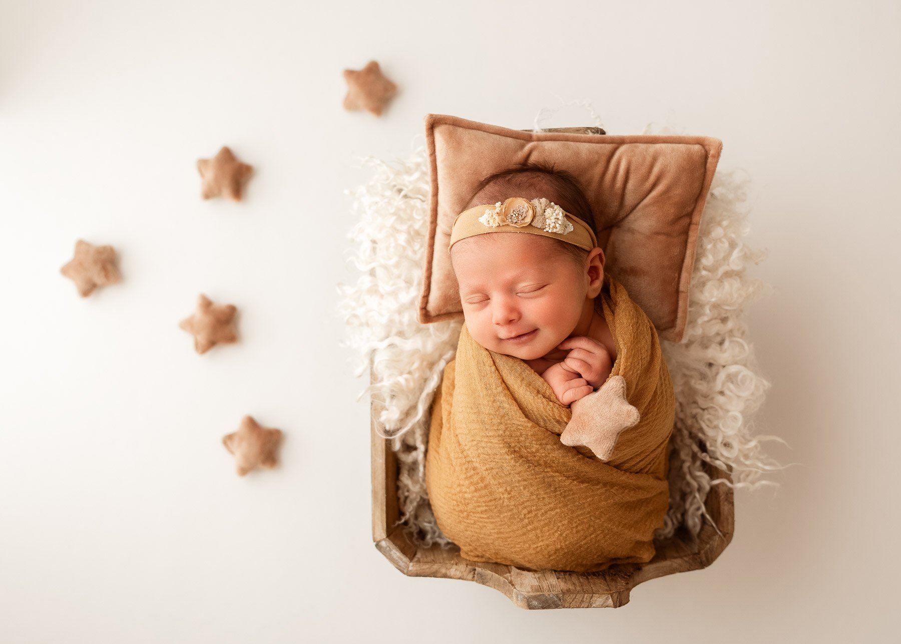 Ein schlafendes neugeborenes Baby in einem braunen Tuch gewickelt, auf einem kleinen Korb liegend, mit einer zarten Blumenkrone auf dem Kopf, und in der Nähe an einem Kinderbett befestigt, umgeben von kleinen Sternen aus Filz auf einem weißen Hinterg