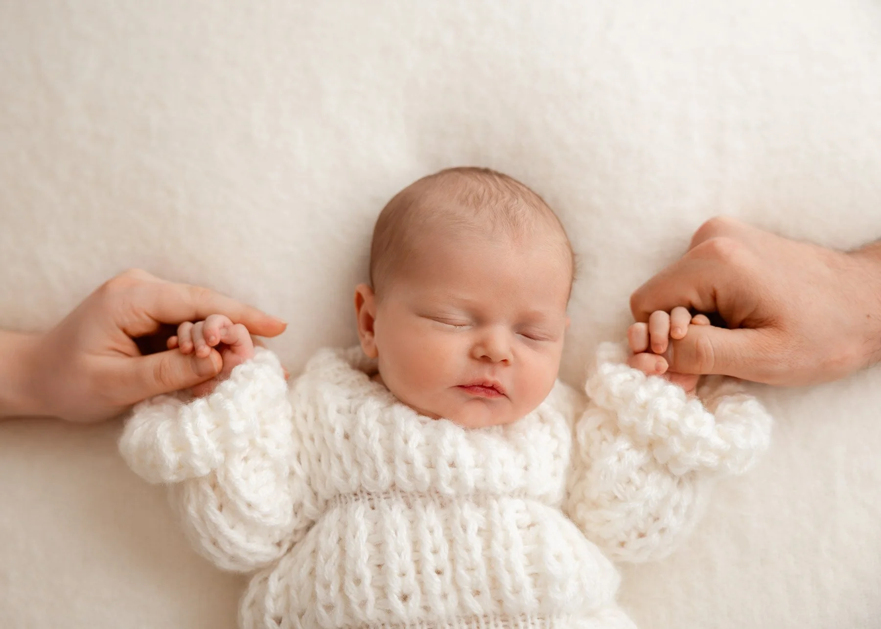 Kleines neugeborenes Baby trägt einen weißen Strickpullover, die Hände von Erwachsenen halten die Hände des Babys, das auf einem weichen, hellen Hintergrund schläft.