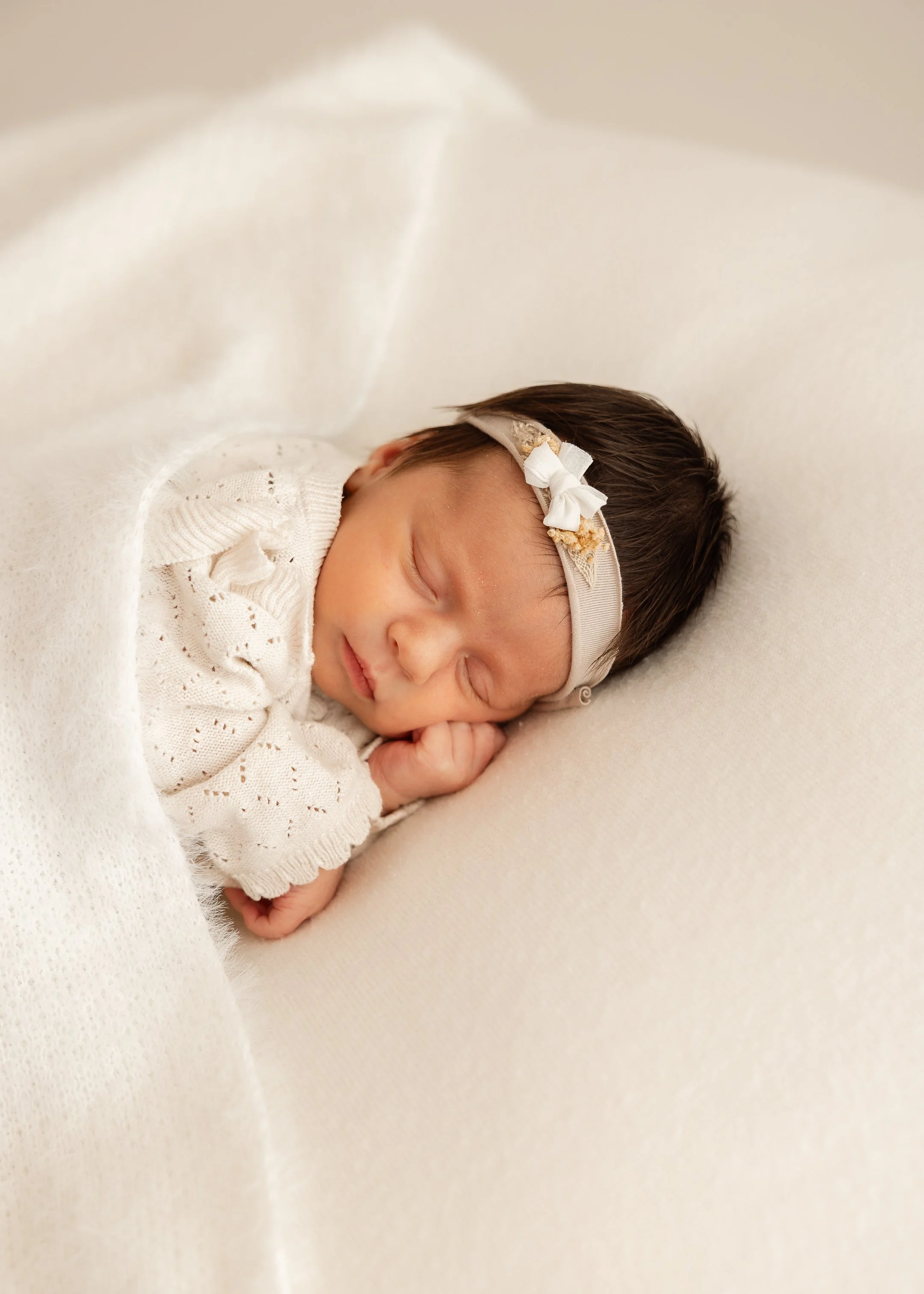 Ein schlafendes Baby mit dunklem Haar, tragend eines weißen Kleidungsstücks, liegt auf einem weichen, hellen Hintergrund und trägt einen Kopfschband mit einer kleinen Schleife.