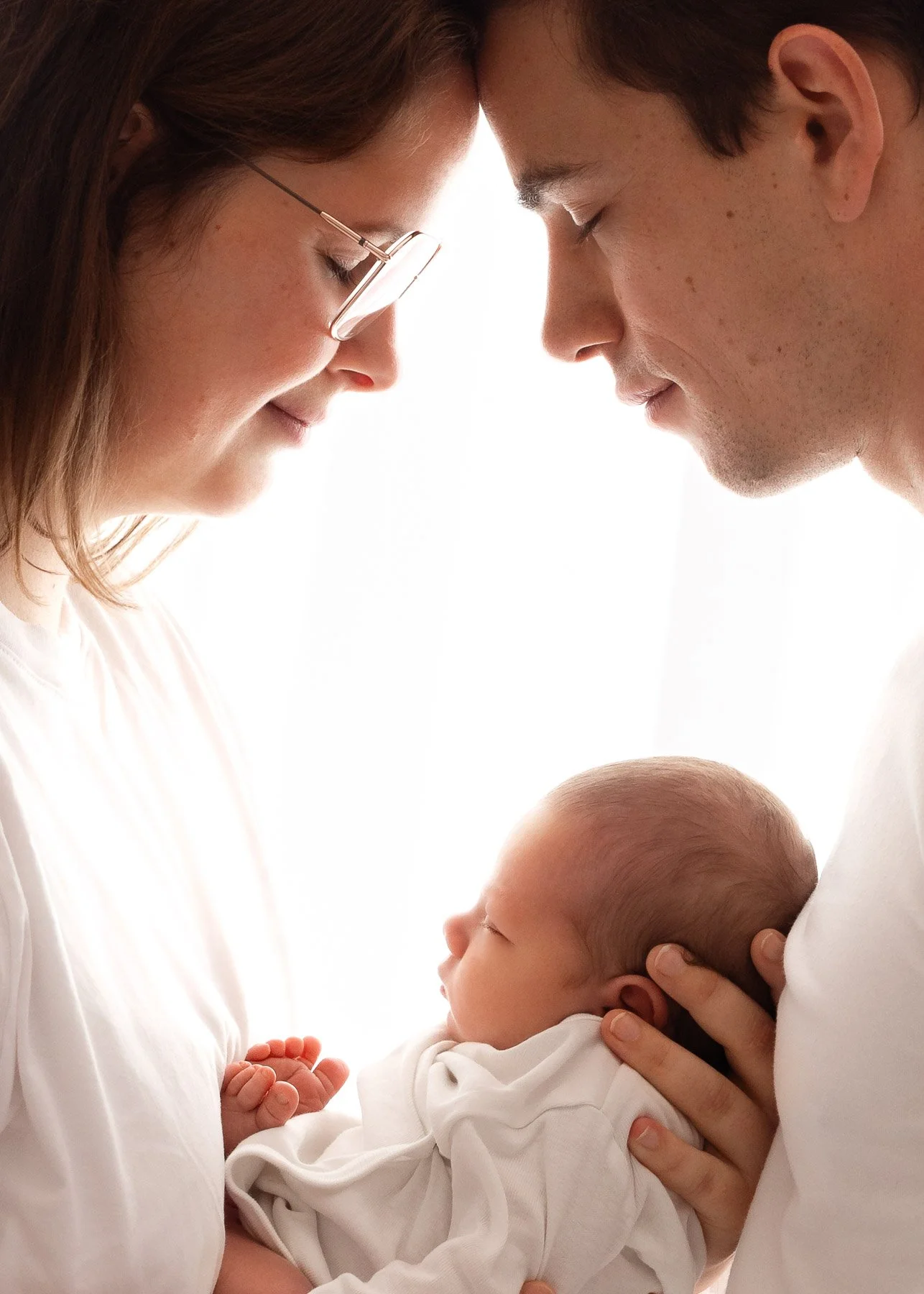 Ein Baby wird von einem Mann und einer Frau gehalten, die beide ihre Stirnen sanft an das des Babys drücken. Die Frau trägt Brille und hat braune Haare, der Mann hat dunkle Haare. Die Szene wirkt liebevoll und intim.
