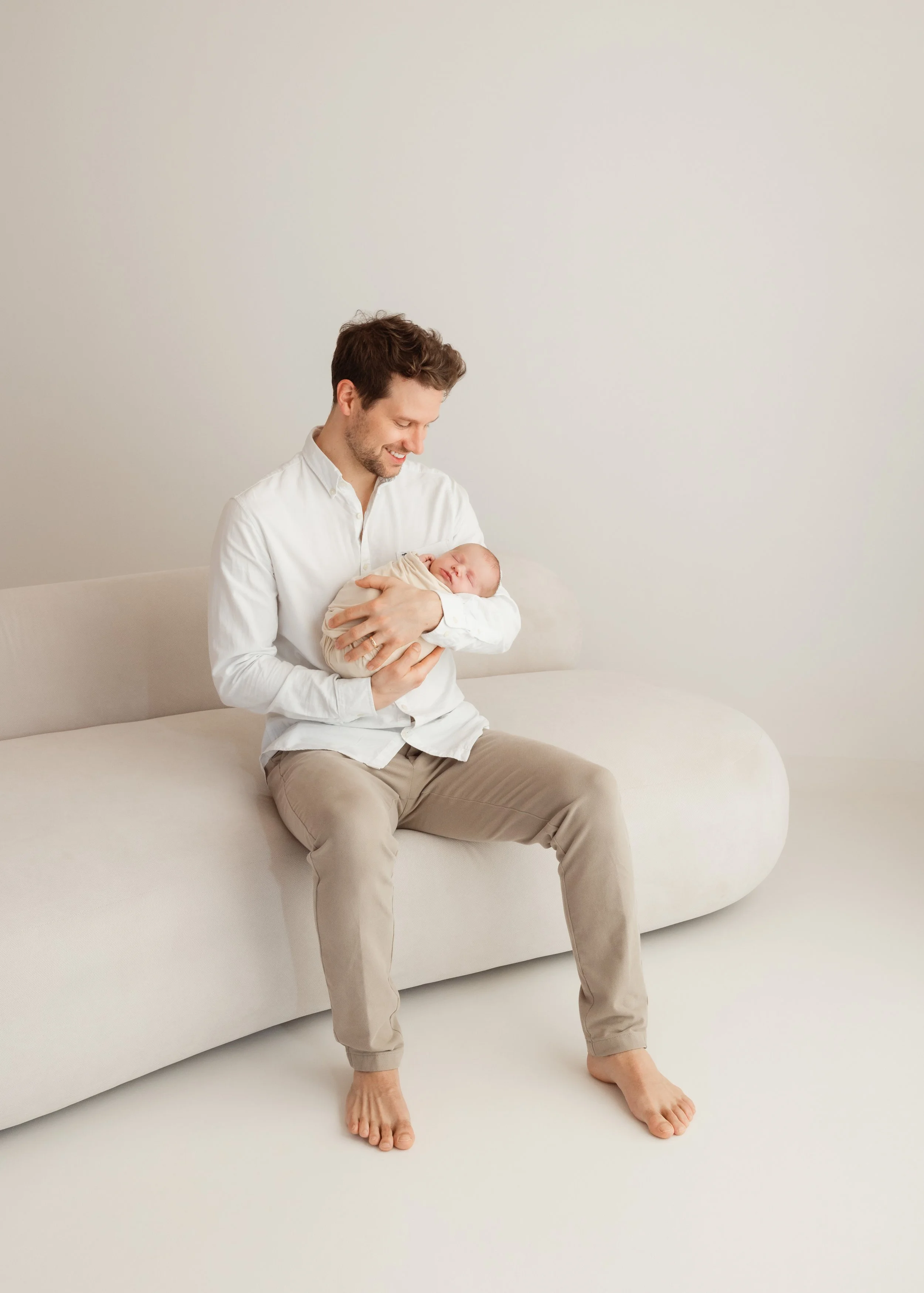 Ein Mann hält ein schlafendes Baby in den Armen, sitzt auf einem weißen Sofa in einem minimalistischen Raum.