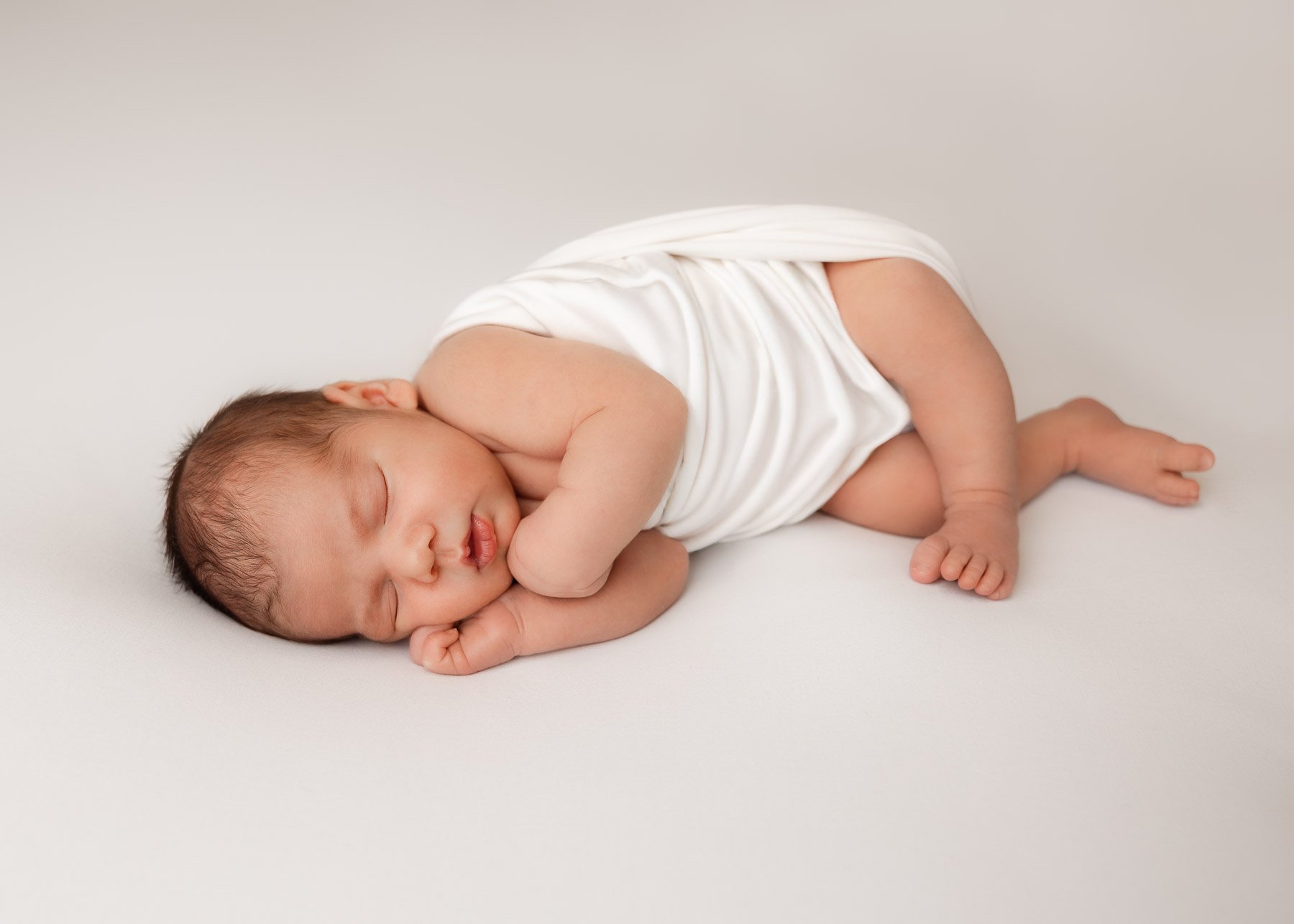 Ein schlafender Säugling liegt auf der Seite, eingekuschelt in einem weißen Tuch, auf einem weißen Hintergrund.