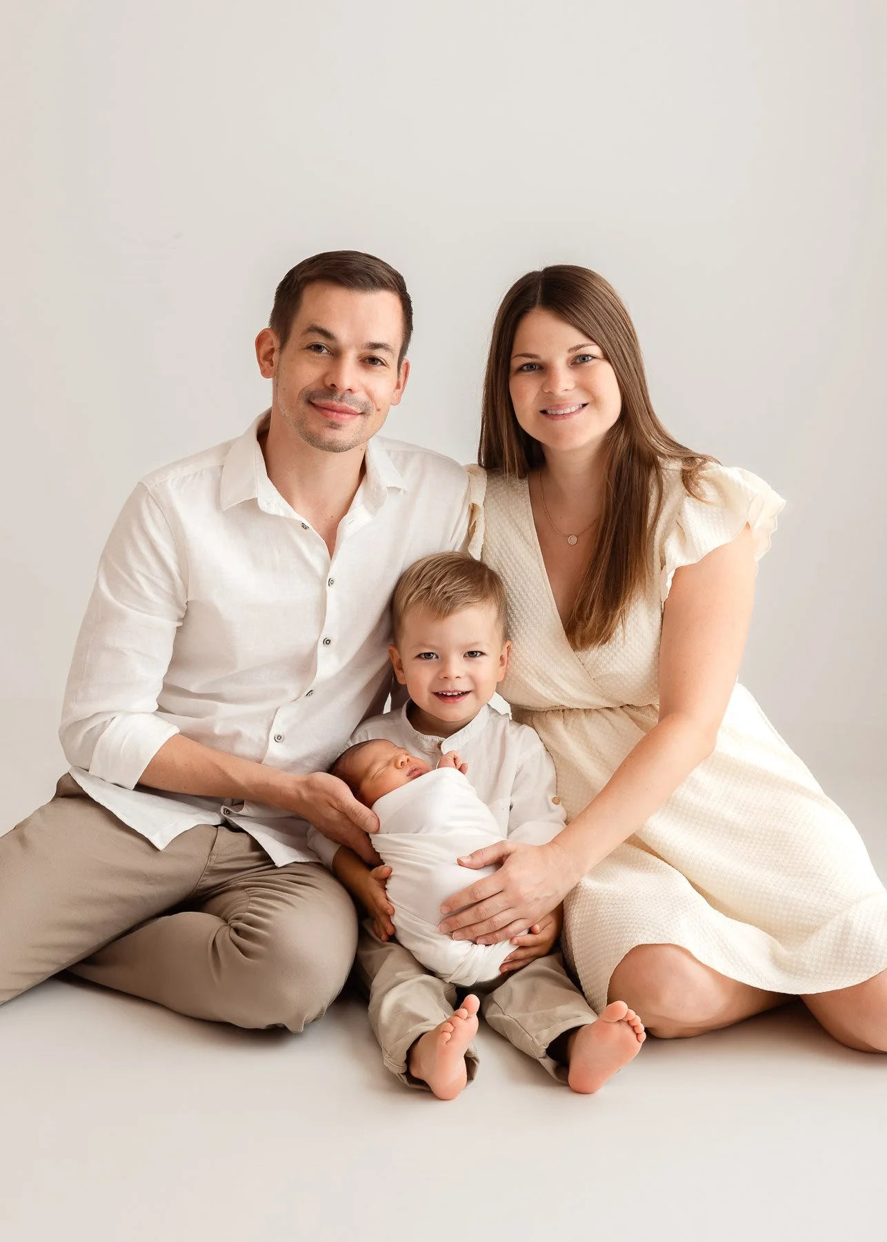 Familie mit vier Mitgliedern, inklusive neugeborenes Baby, auf einem weißen Hintergrund, sitzend und lachend.