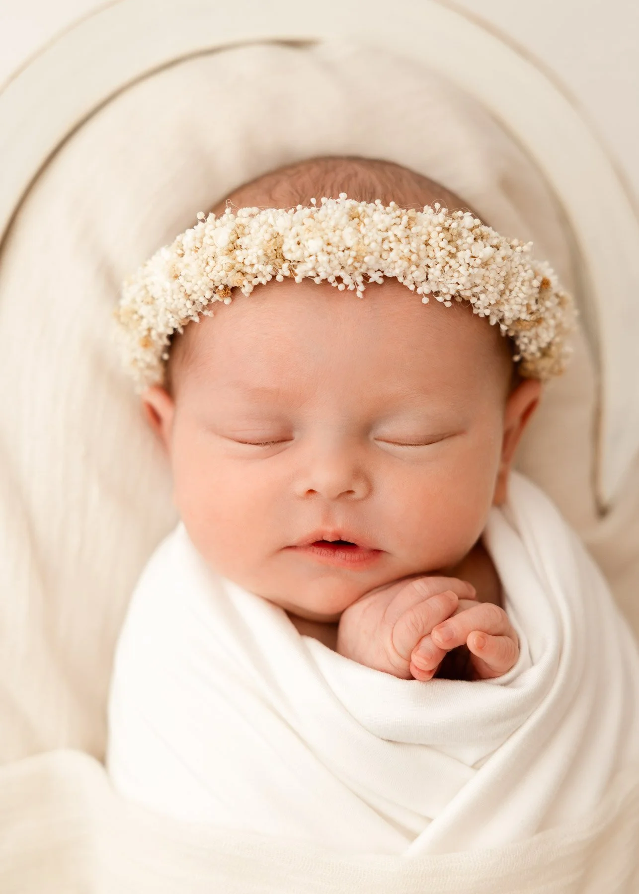 Ein schlafendes Baby mit einem Blütenkranz auf dem Kopf, in weißem Tuch eingewickelt, liegt auf einer weichen, hellen Unterlage.