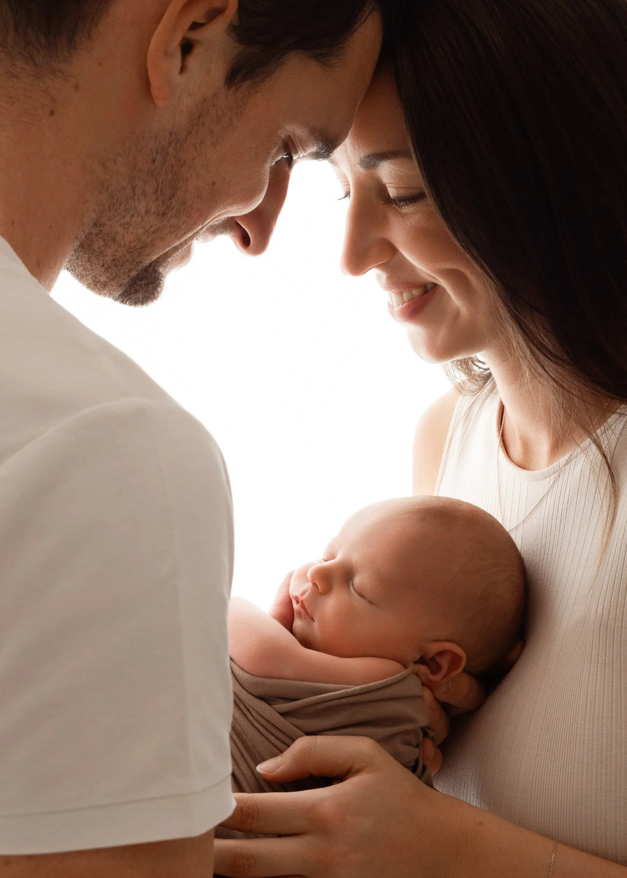 Ein Paar hält ein schlafendes Baby liebevoll im Arm, beide blicken sich zärtlich an, im Hintergrund helles Licht.