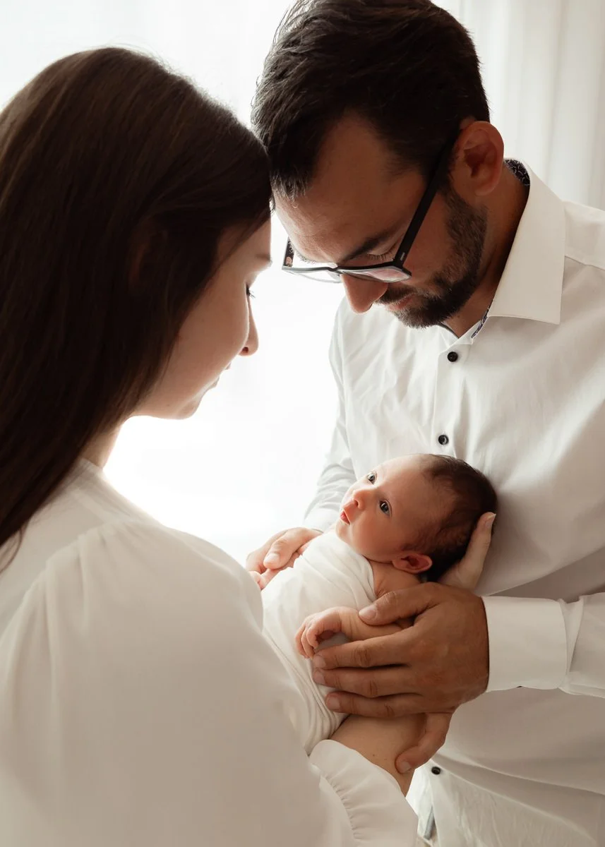 Ein Paar in weißer Kleidung hält ein neugeborenes Baby in den Armen und schaut liebevoll darauf.