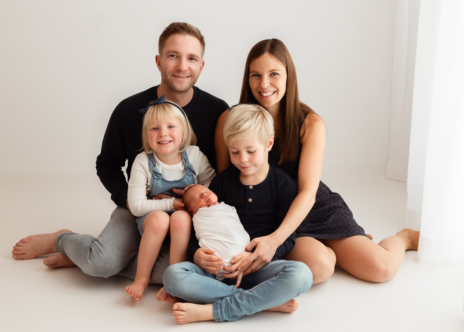 Familie mit vier Erwachsenen und drei Kindern sitzt auf einem weißen Boden in einem hellen Raum, um ein neugeborenes Baby, alle lachen und schauen in die Kamera.