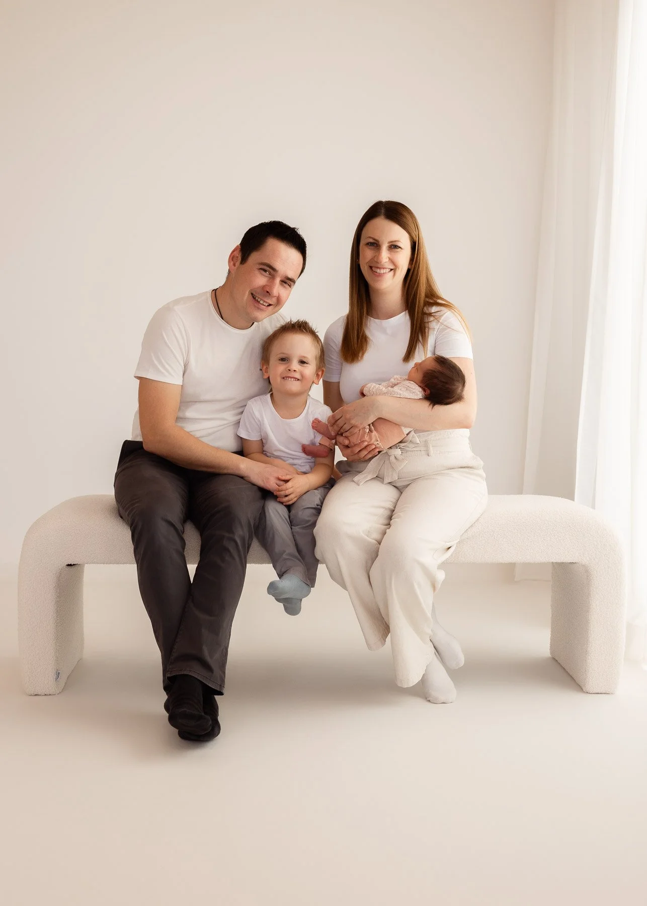 Familienporträt mit vier Personen: zwei Erwachsene, ein Kleinkind und ein Baby, die auf einer modernen weißen Bank sitzen. Alle tragen helle Kleidung vor einem neutralen Hintergrund.