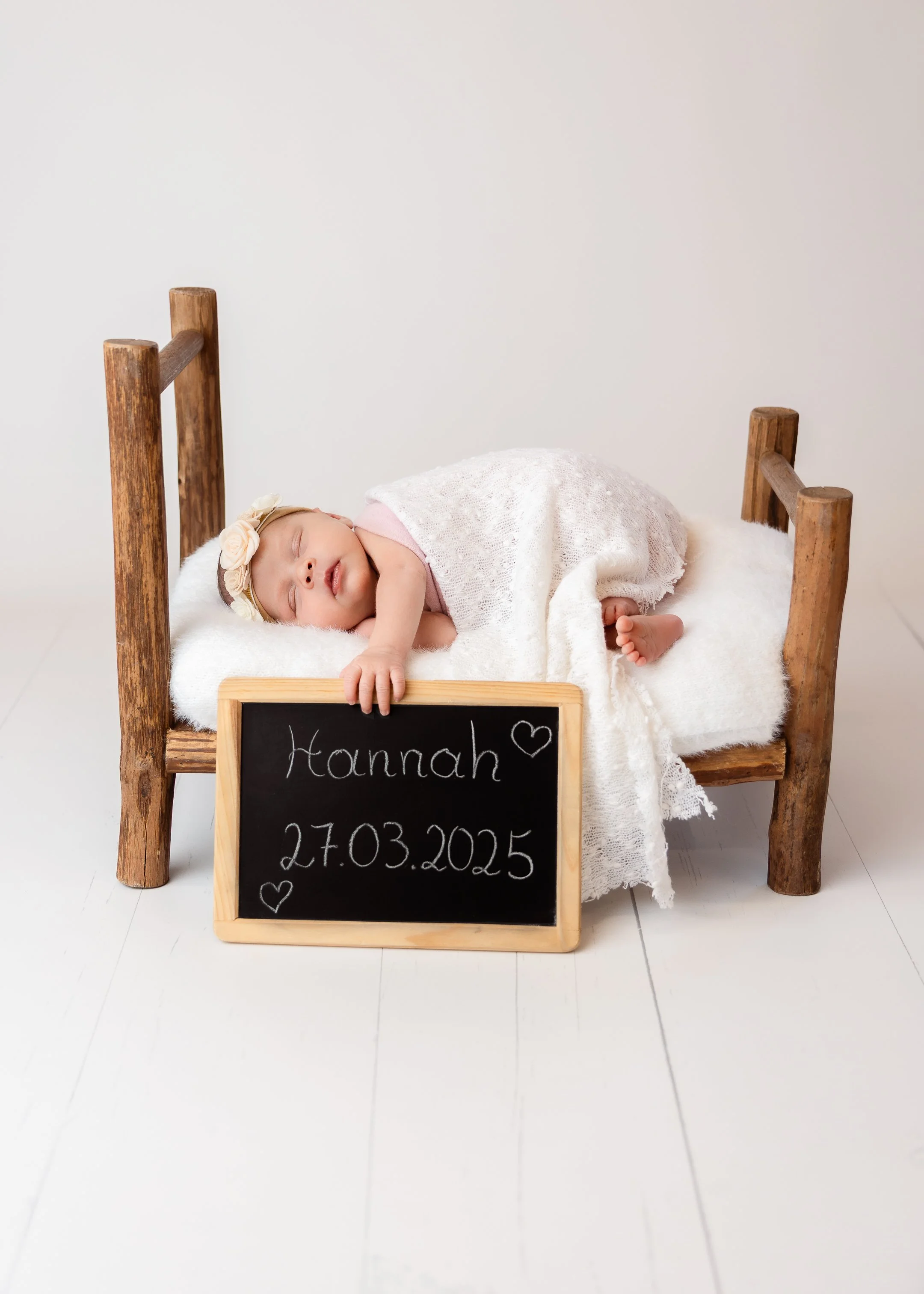 Ein schlafendes Baby liegt auf einem kleinen Holzbett, bedeckt mit einem weißen Tuch, neben einem schwarzen Tafel mit dem Namen Hannah und dem Geburtsdatum 27.03.2025, verziert mit kleinen Herzen.