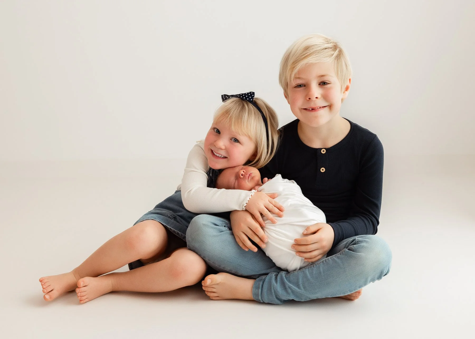 Drei Kinder, ein Mädchen, ein Junge und ein Neugeborenes, sitzen gemeinsam auf dem Boden vor hellem Hintergrund. Das Mädchen hält das schlafende Neugeborene, die beiden älteren Kinder lächeln.