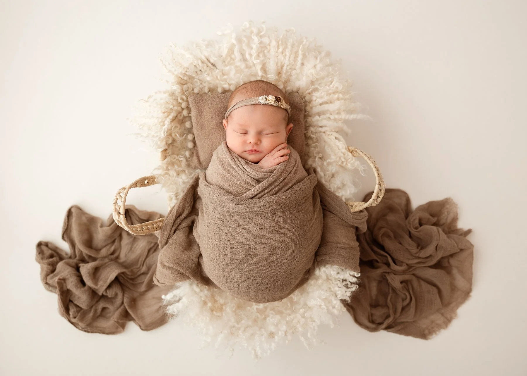 Ein schlafender Säugling, eingewickelt in braunen Stoff, liegt auf einem weißen, lockigen Decke- und Kissen-Arrangement.