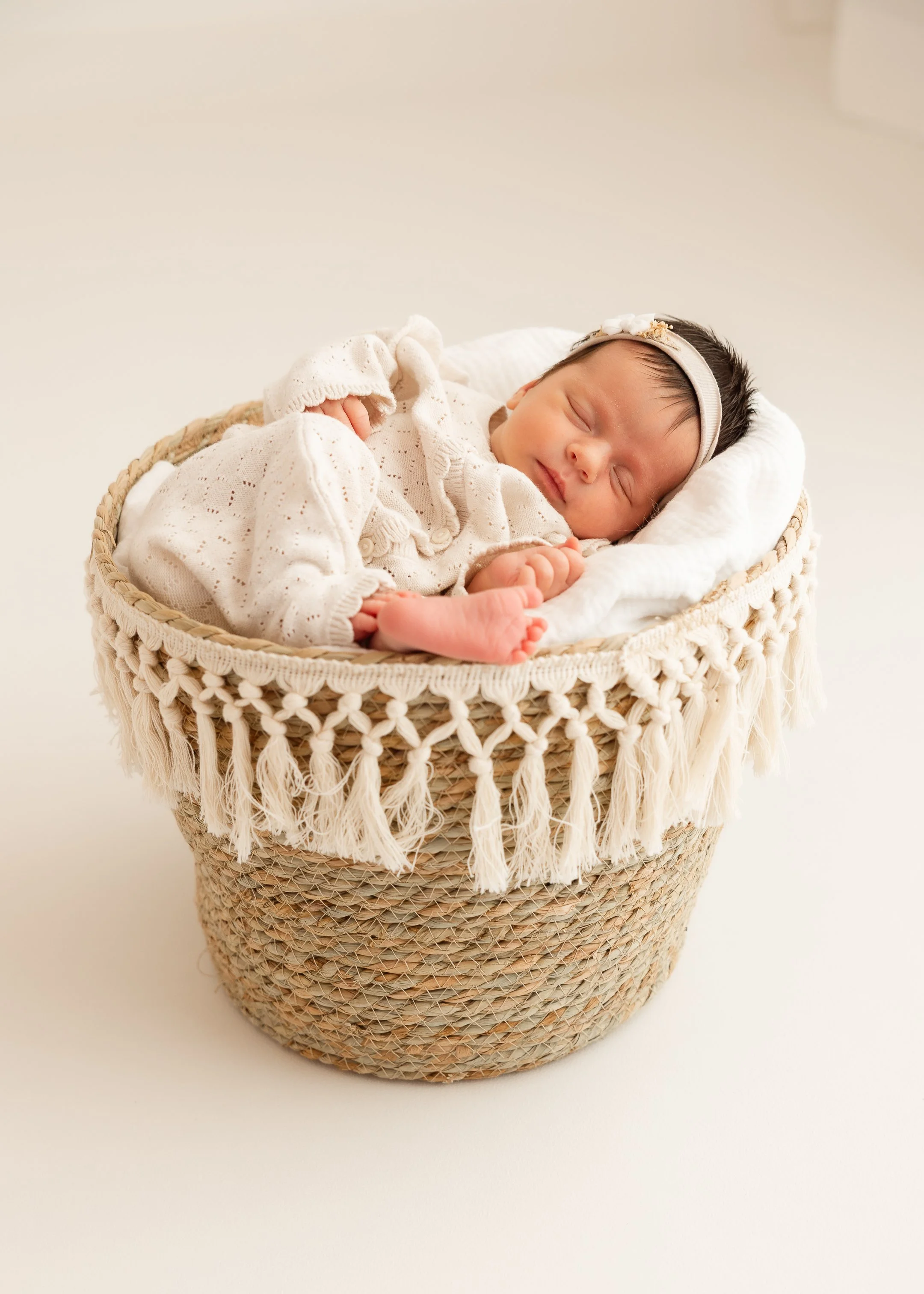 Ein schlafendes Baby in einem Korb mit einem weißen Tuch, getragen von einem weichen, cremefarbenen Kleid und einem weißen Kopfband.